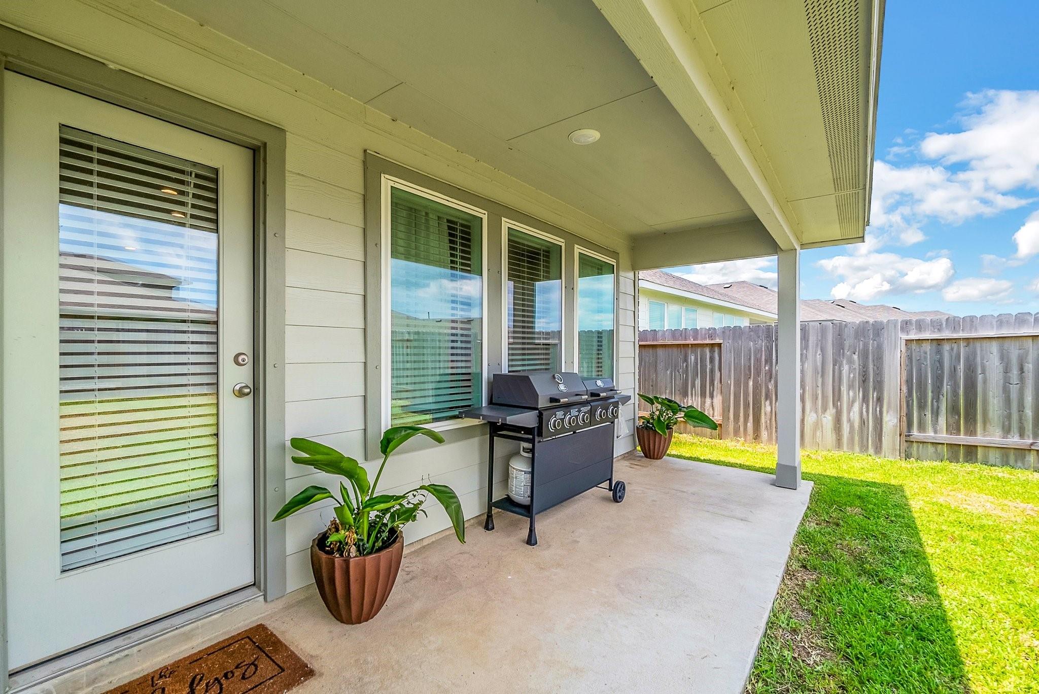 Covered backyard patio with a barbecue grill, potted plants, and a fenced lawn.