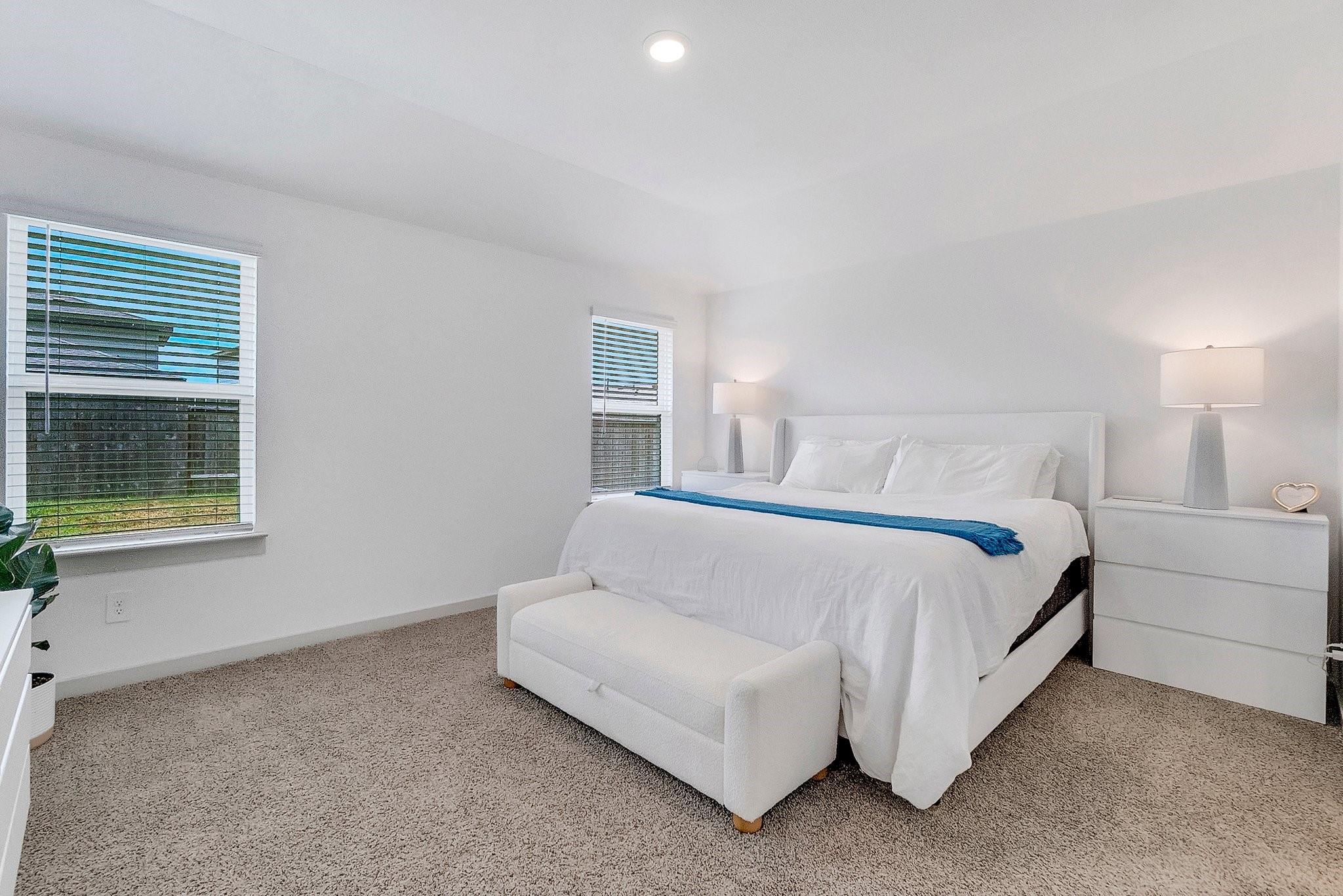 Bedroom with a white bed, upholstered bench, gray carpet, and two windows with blinds.
