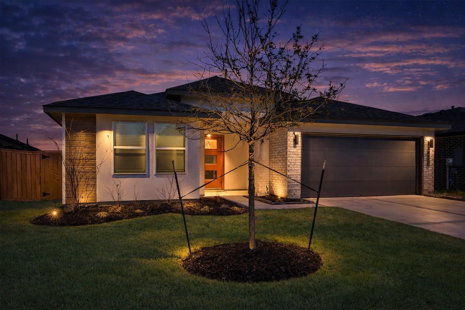 Single-story home at dusk with a lit entry, two-car garage, and young tree in the front lawn.
