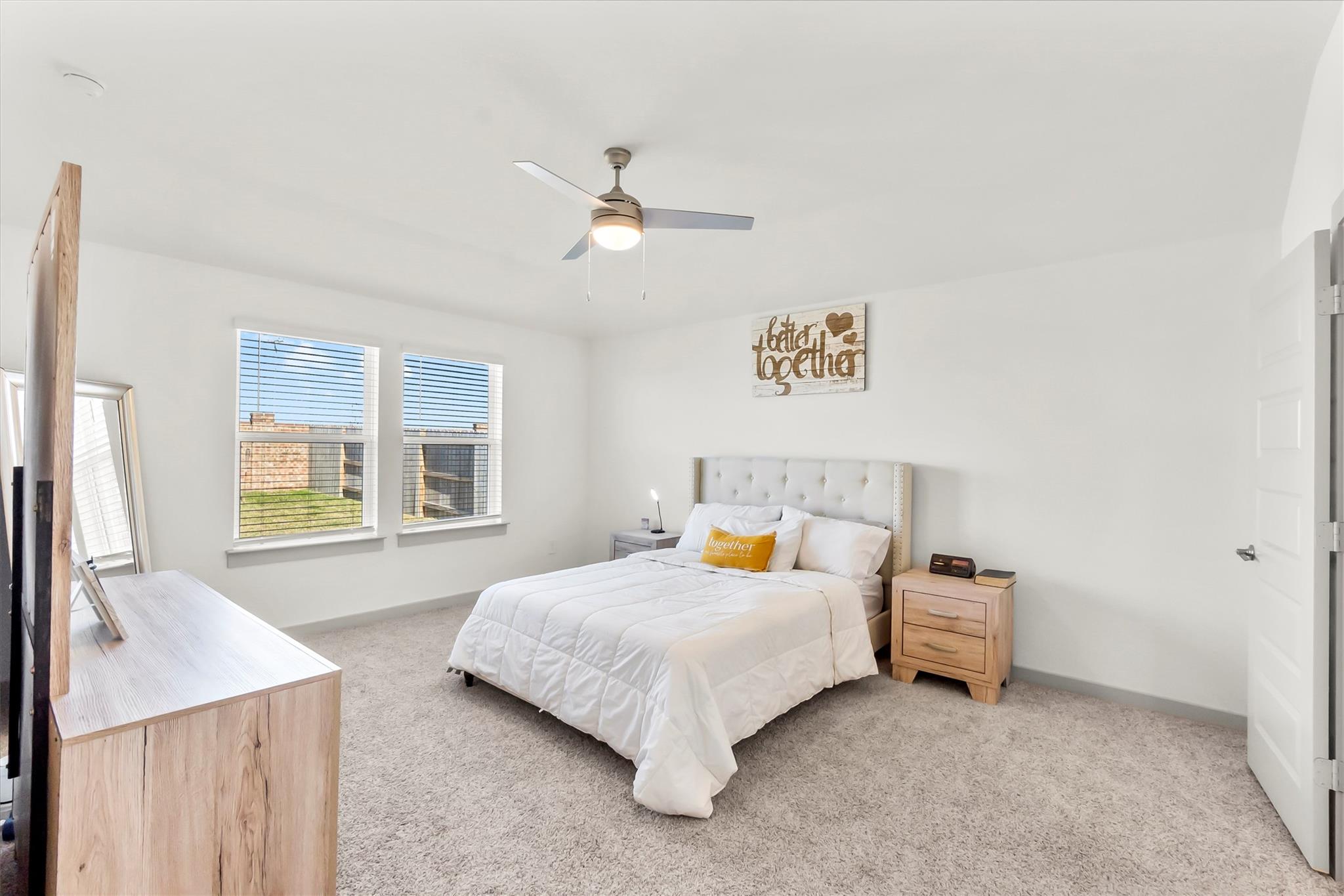 Staged bedroom with a white bed, tufted headboard, two windows, and light wood nightstand.