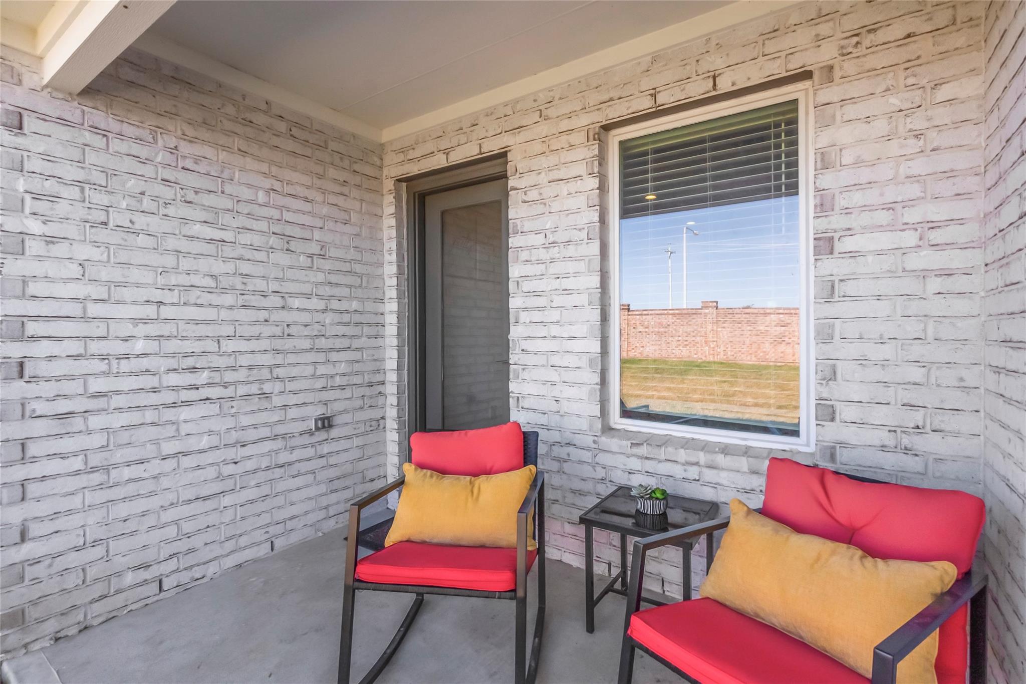 Covered white-brick porch with red-cushioned chairs, yellow pillows, and a small table.