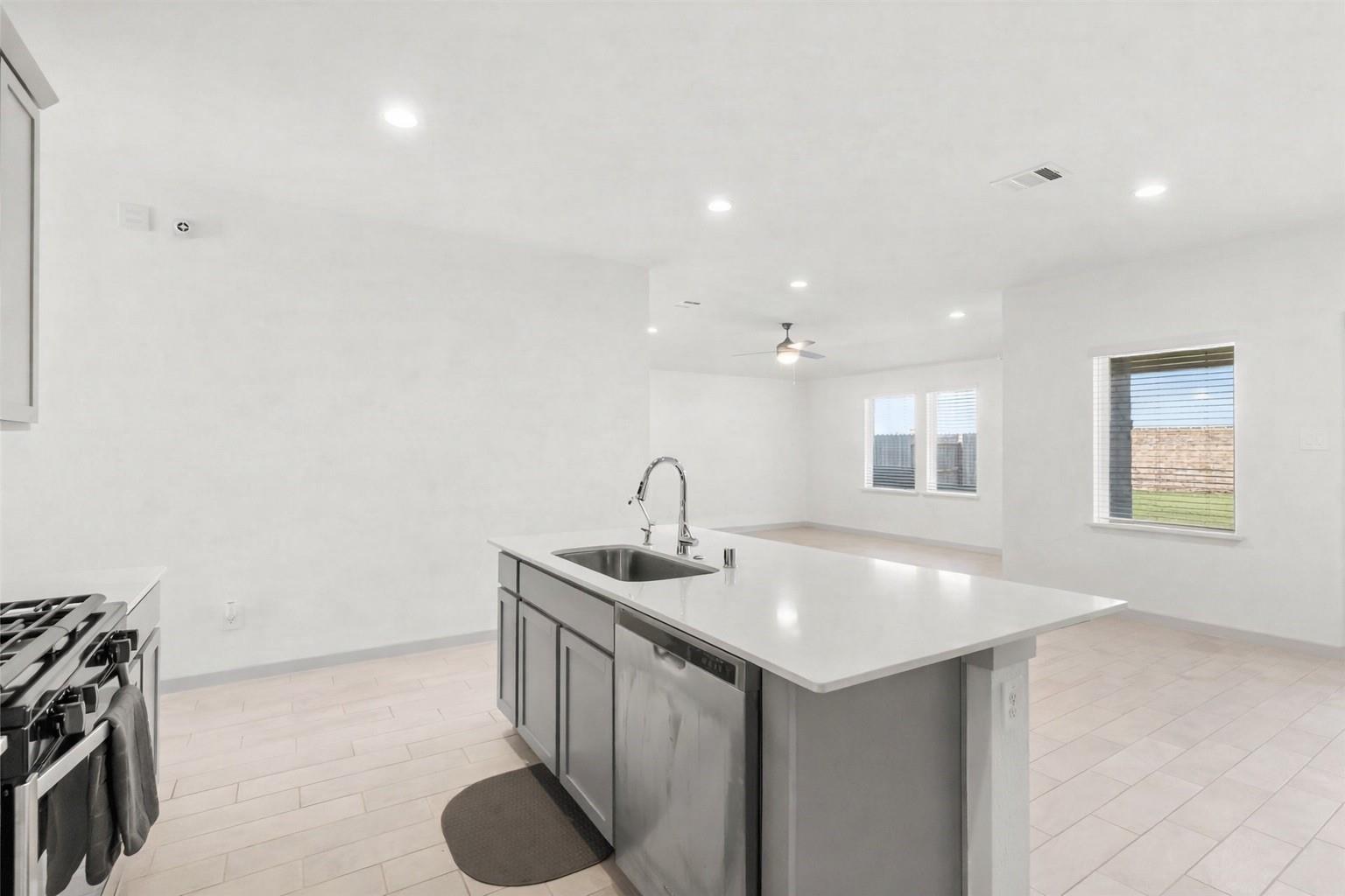 Bright kitchen with a large white island, sink, dishwasher, and partially visible stainless-steel gas range.