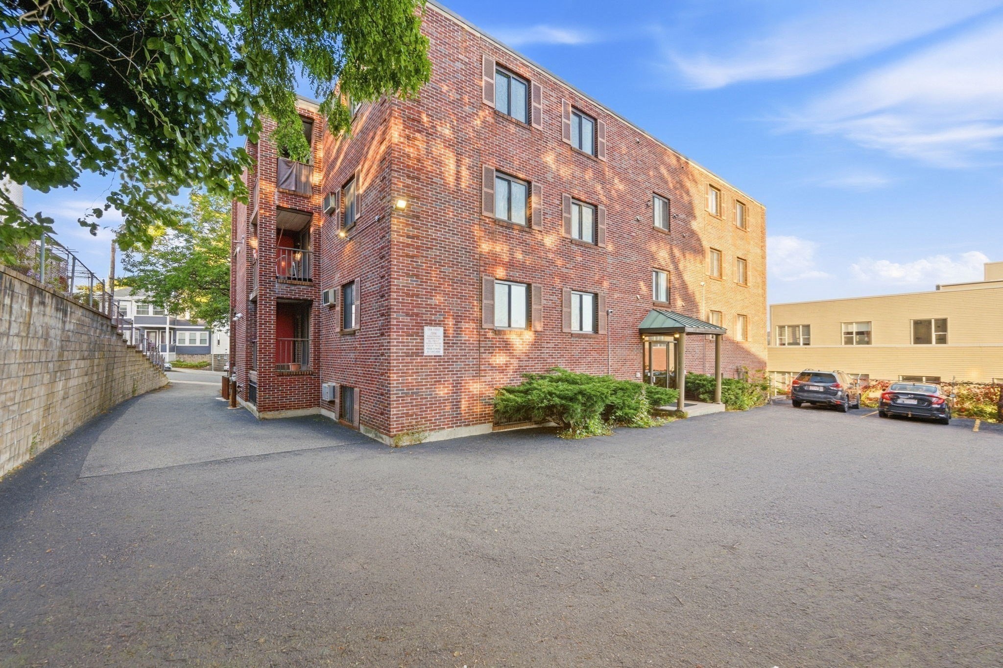 Red brick apartment building with a covered entrance and paved parking area.