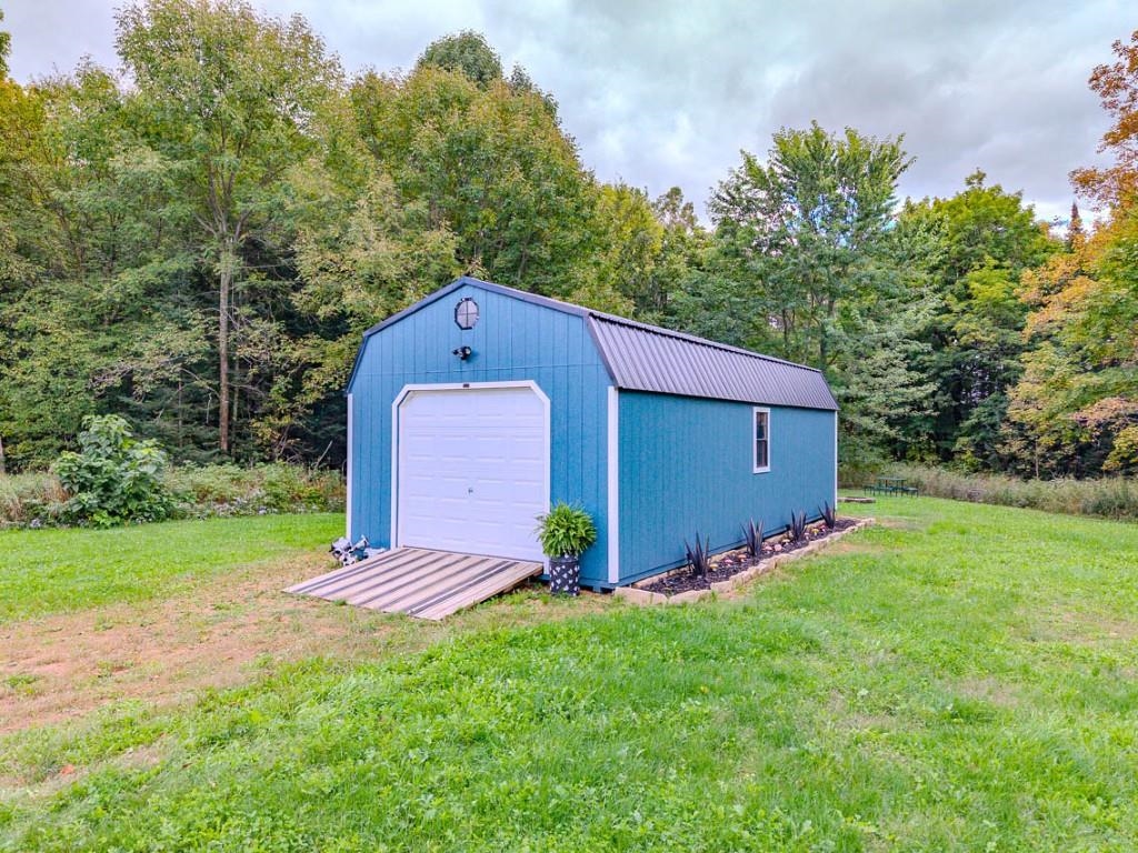 Blue detached shed with a white garage door and wooden ramp in a wooded clearing.