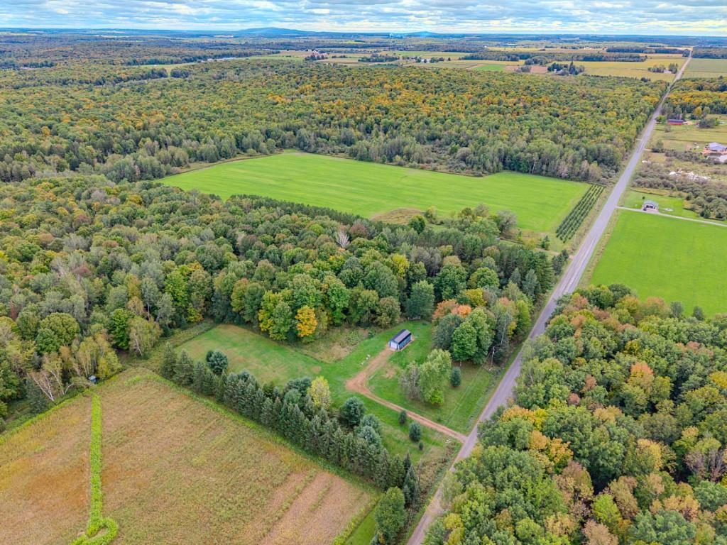 Aerial view of wooded rural acreage with open fields, a small outbuilding, and a road.