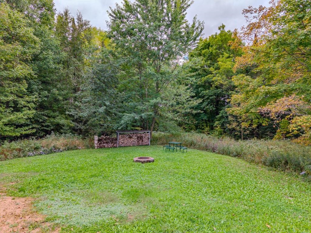 Wooded backyard with a grassy lawn, circular fire pit, woodpile, and small outdoor table.