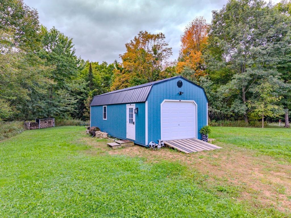 Blue detached shed with a white garage door and wooden ramp, surrounded by autumn trees.