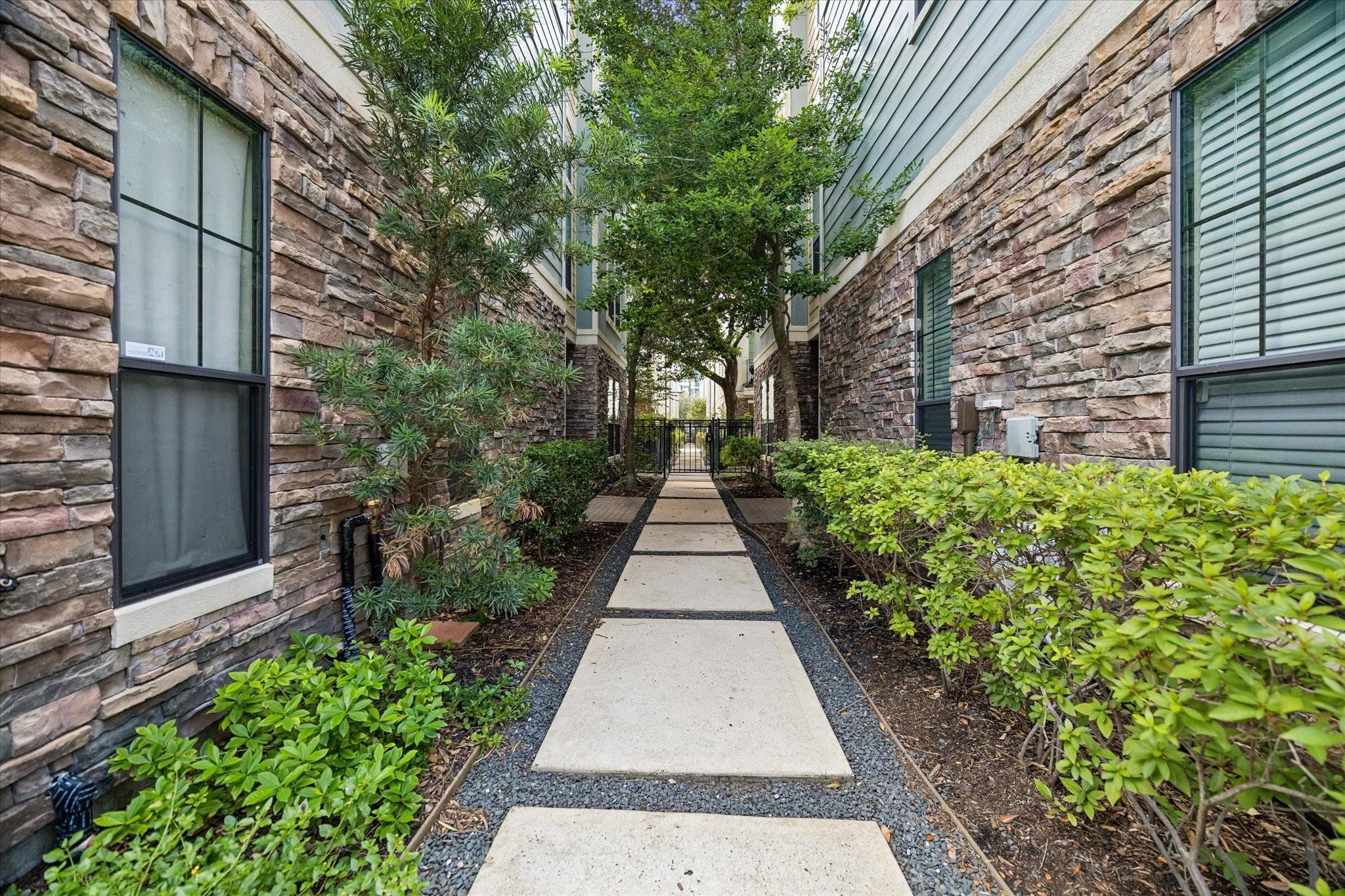 Landscaped walkway between stone-clad buildings, with concrete stepping slabs leading to a gate.