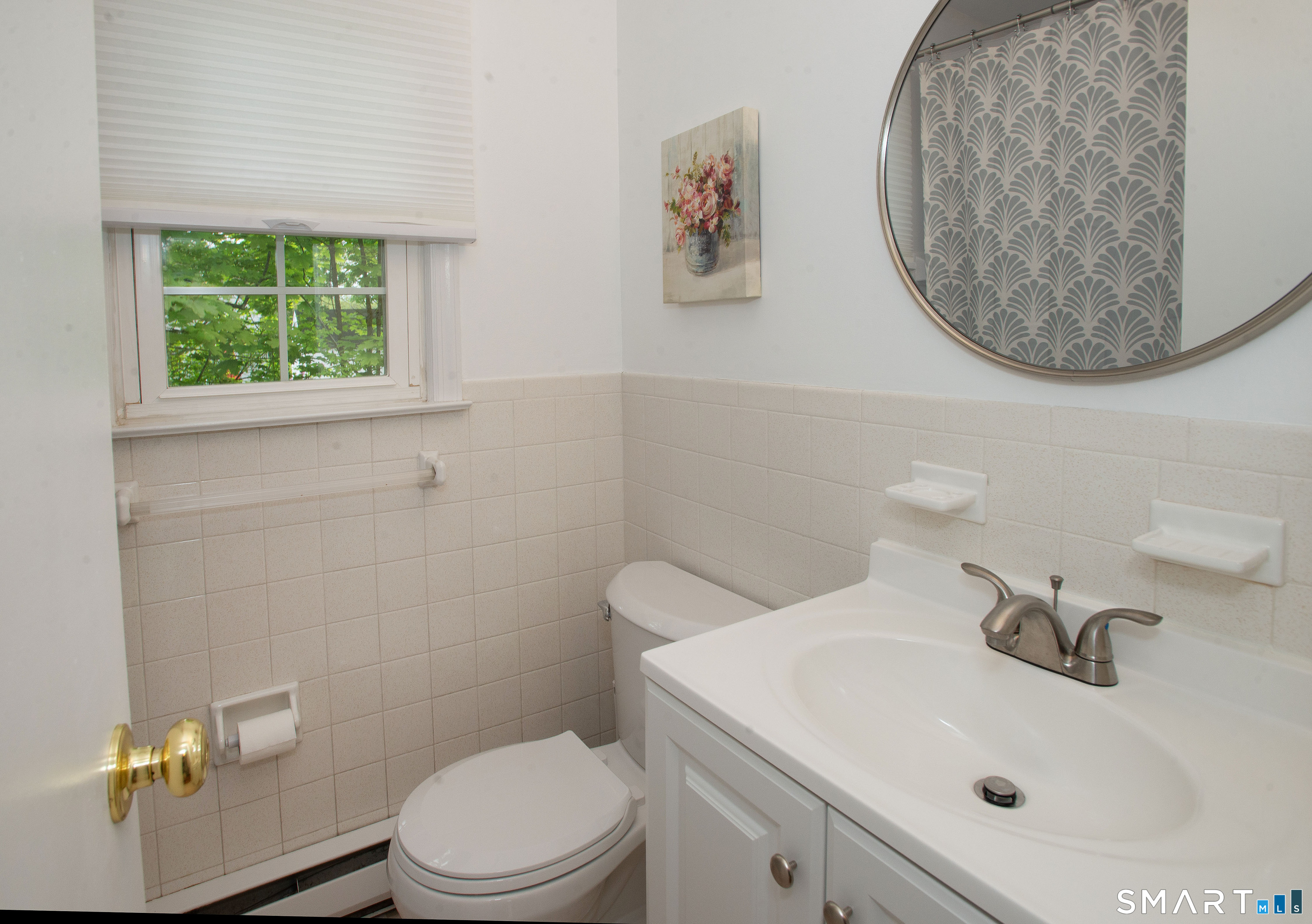 Bathroom with a white vanity, toilet, beige tile, and round mirror beside a leafy window.