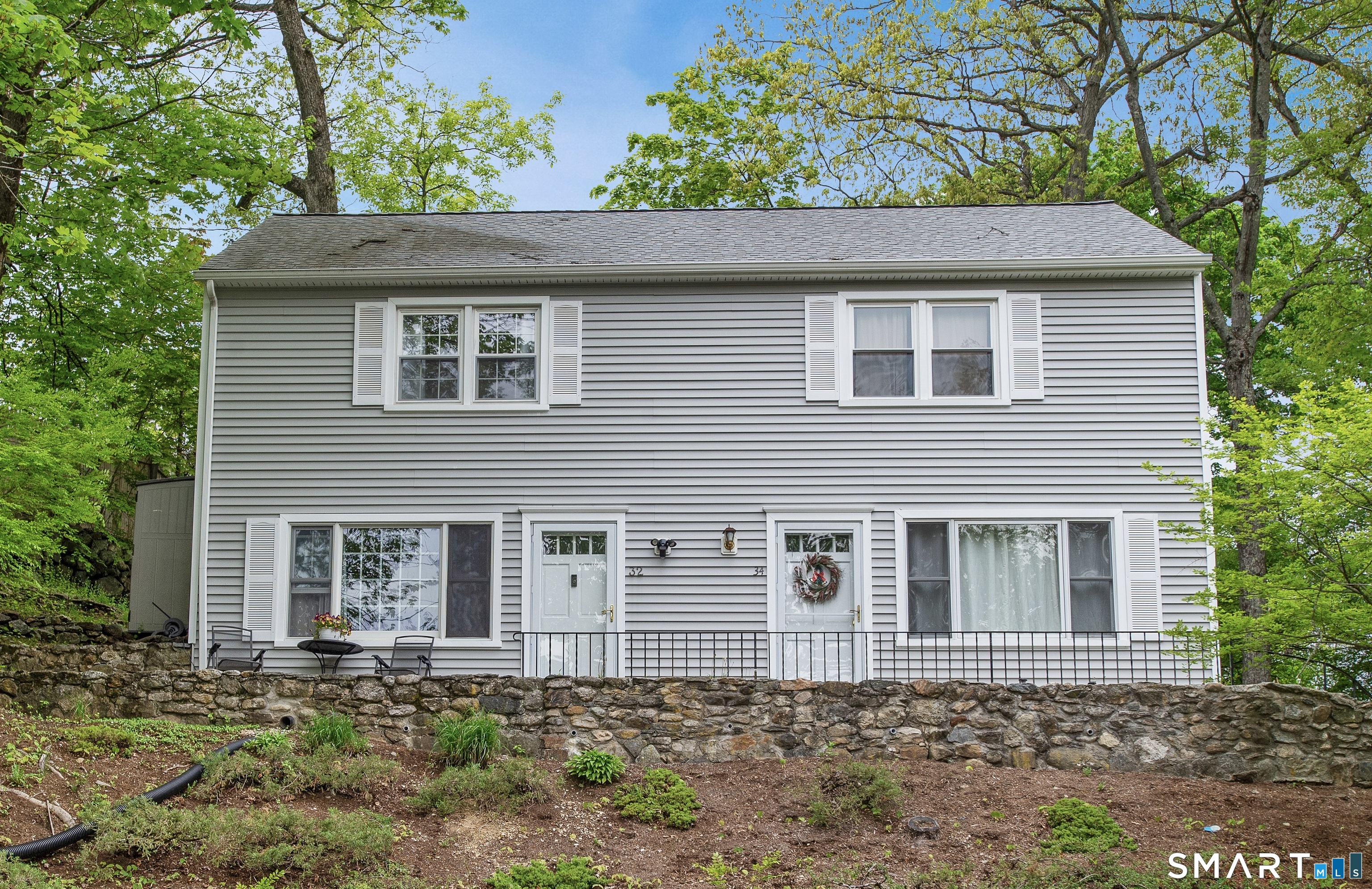 Gray two-story residence with two entrances, white shutters, a stone retaining wall, and leafy trees.