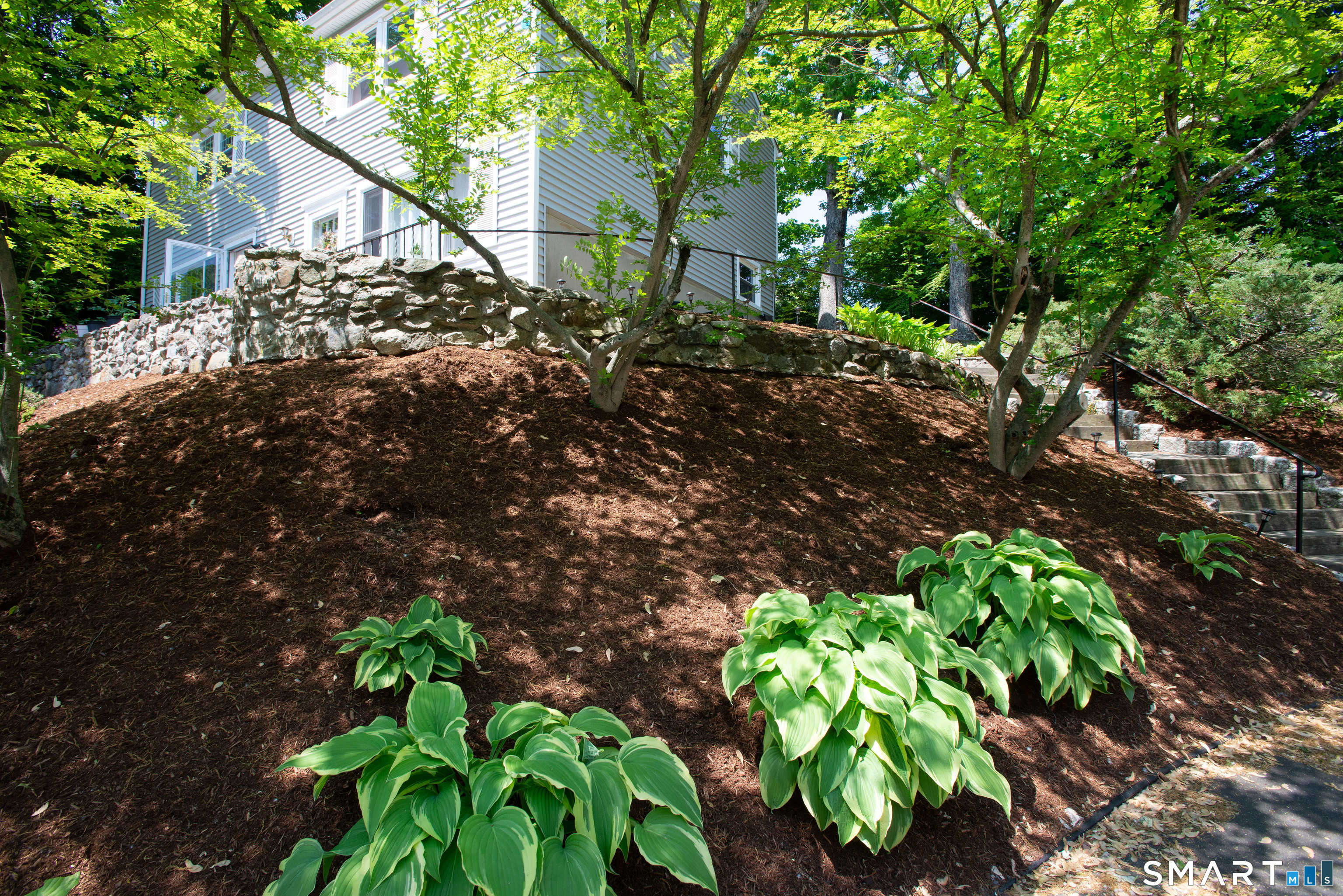 White-sided home above a stone retaining wall, framed by mature trees and landscaped hostas.