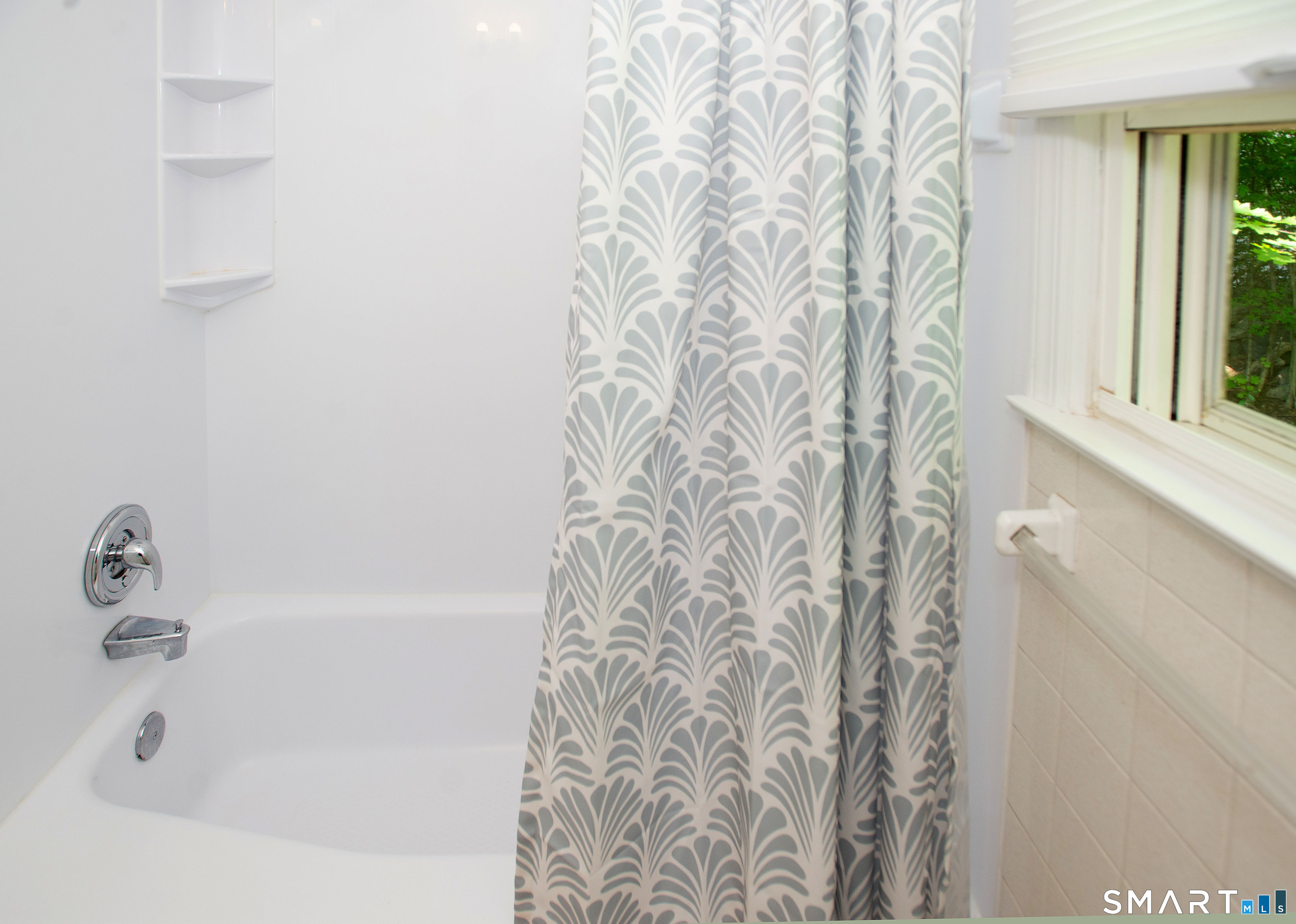 Bathroom tub with chrome fixtures, patterned shower curtain, and a window overlooking greenery.