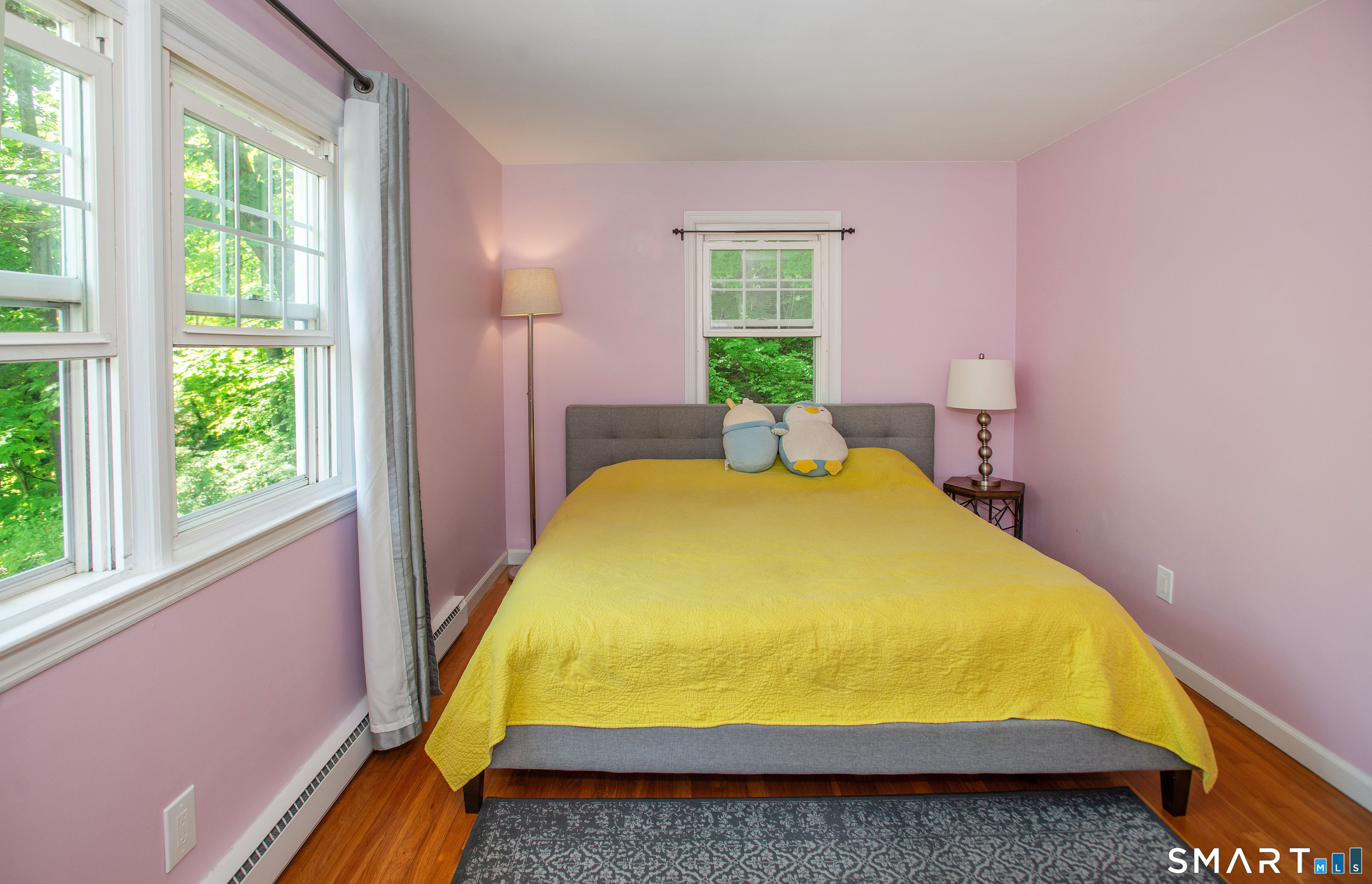 Bedroom with pink walls, yellow quilt, plush toys, and large windows overlooking greenery.