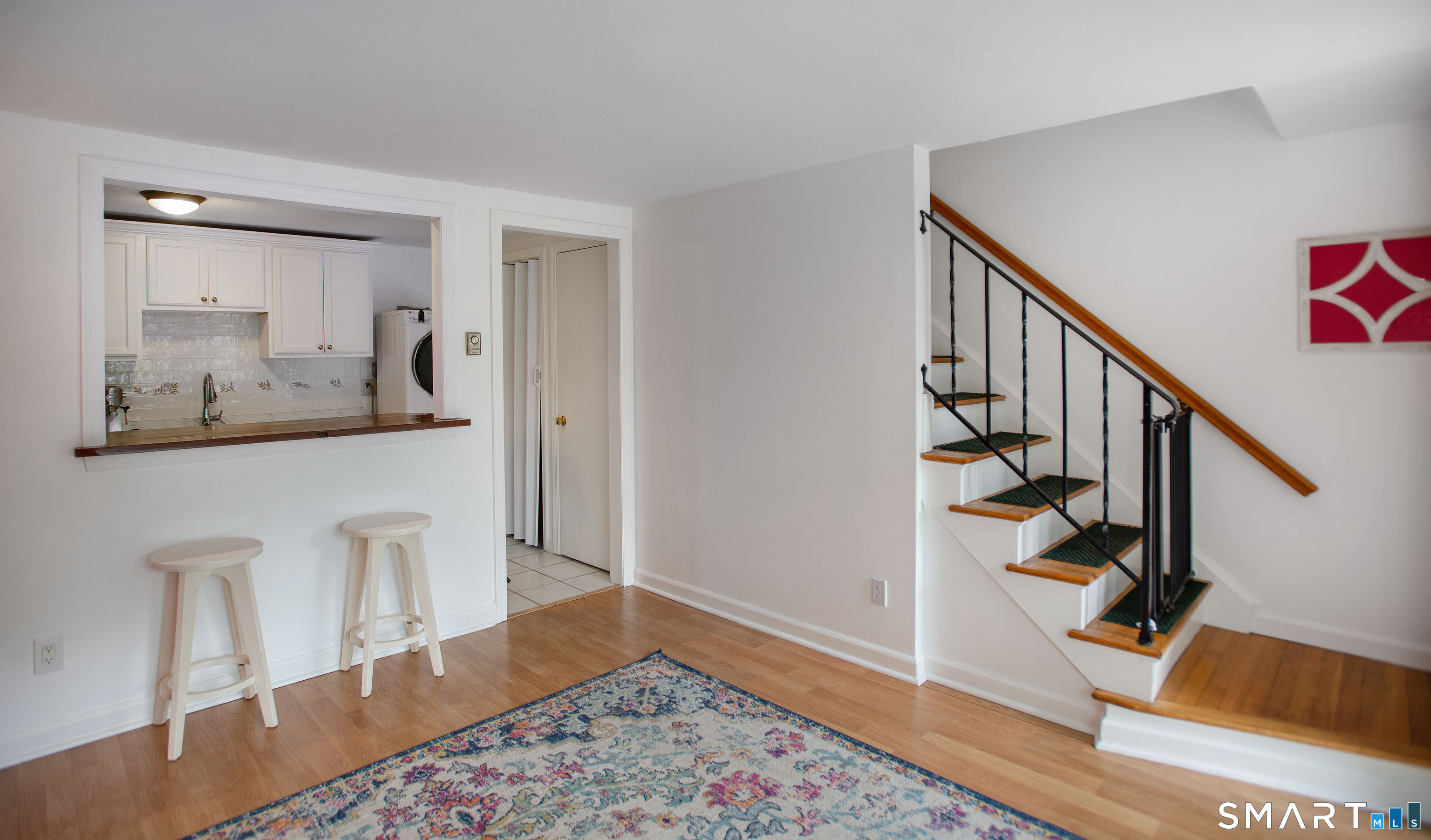 Entry area with a wood staircase, kitchen pass-through, two stools, and patterned rug.