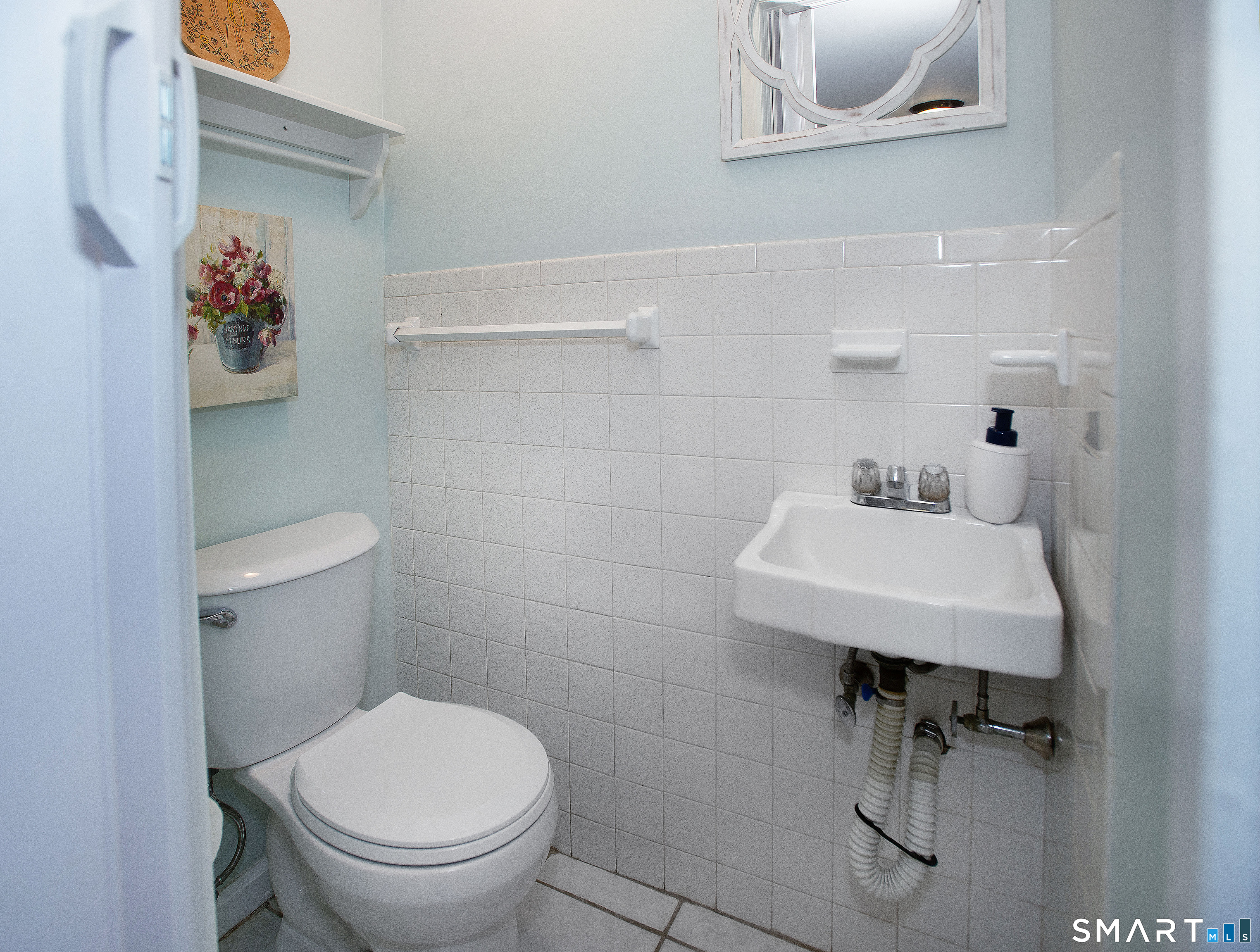 Compact tiled bathroom with a white toilet, wall-mounted sink, grab bars, and floral wall art.