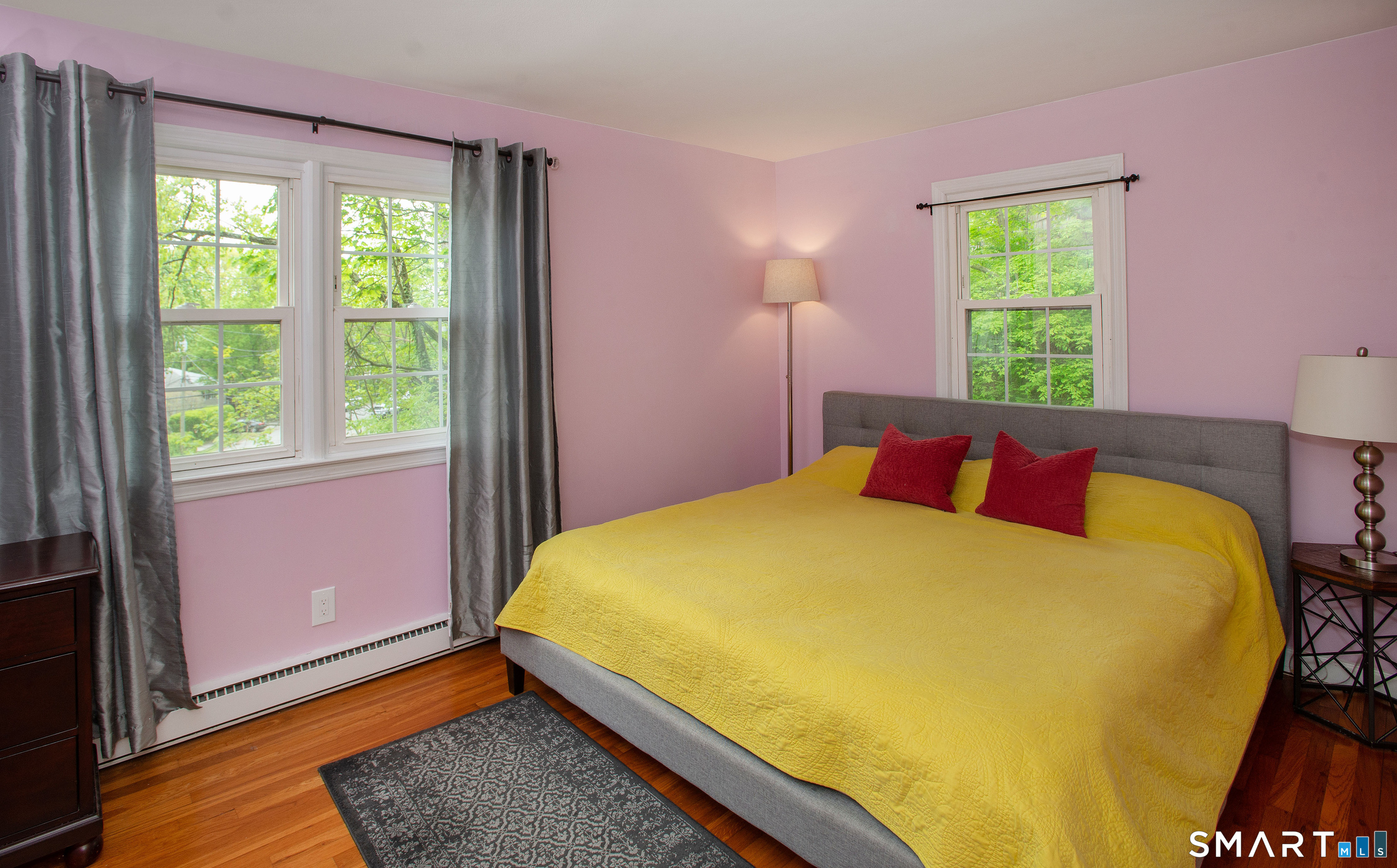 Bedroom with pink walls, a yellow quilt, red pillows, hardwood floors, and three windows overlooking trees.