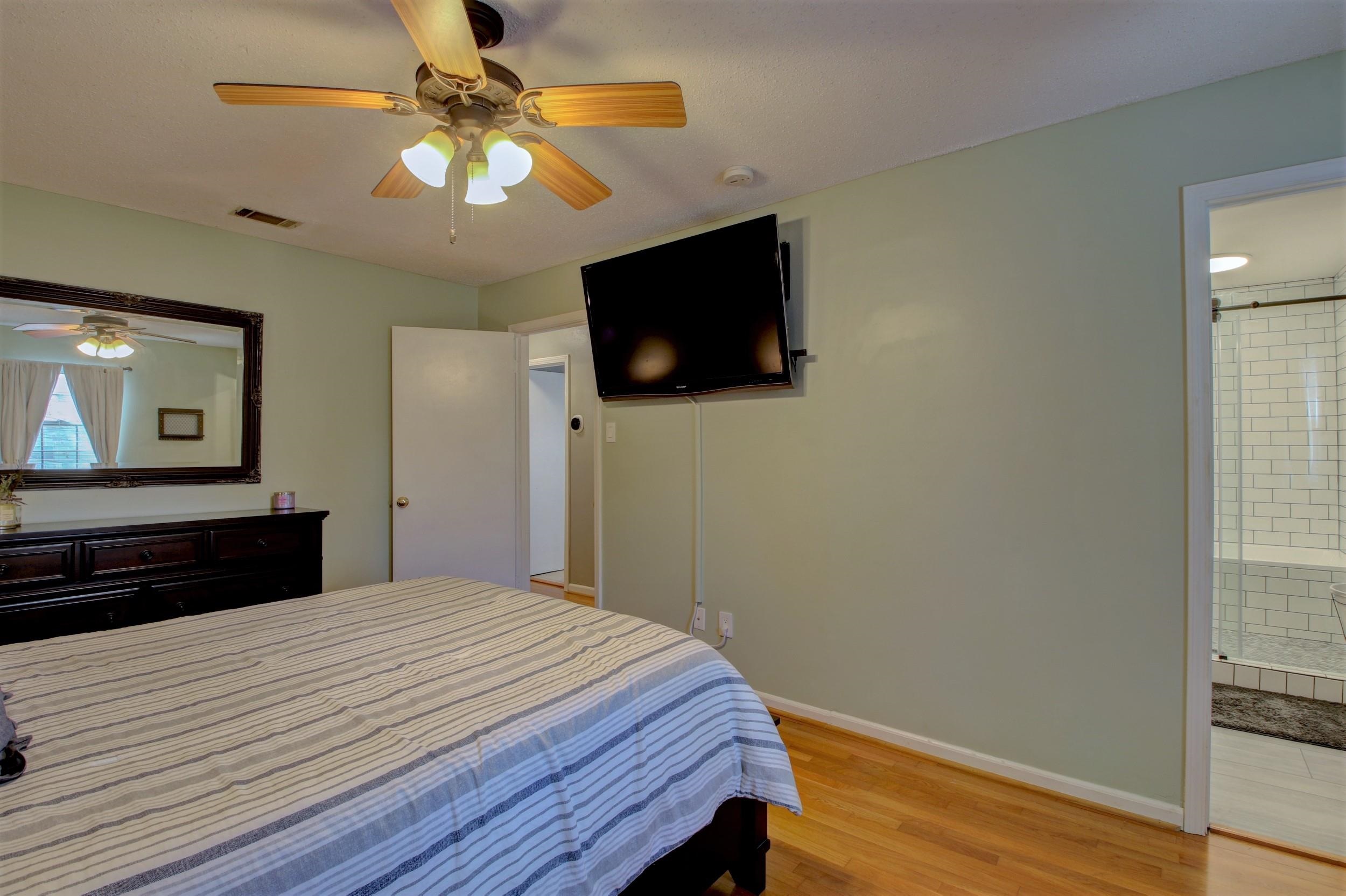 Bedroom with striped bedding, a ceiling fan, wall-mounted TV, and an adjoining tiled shower.