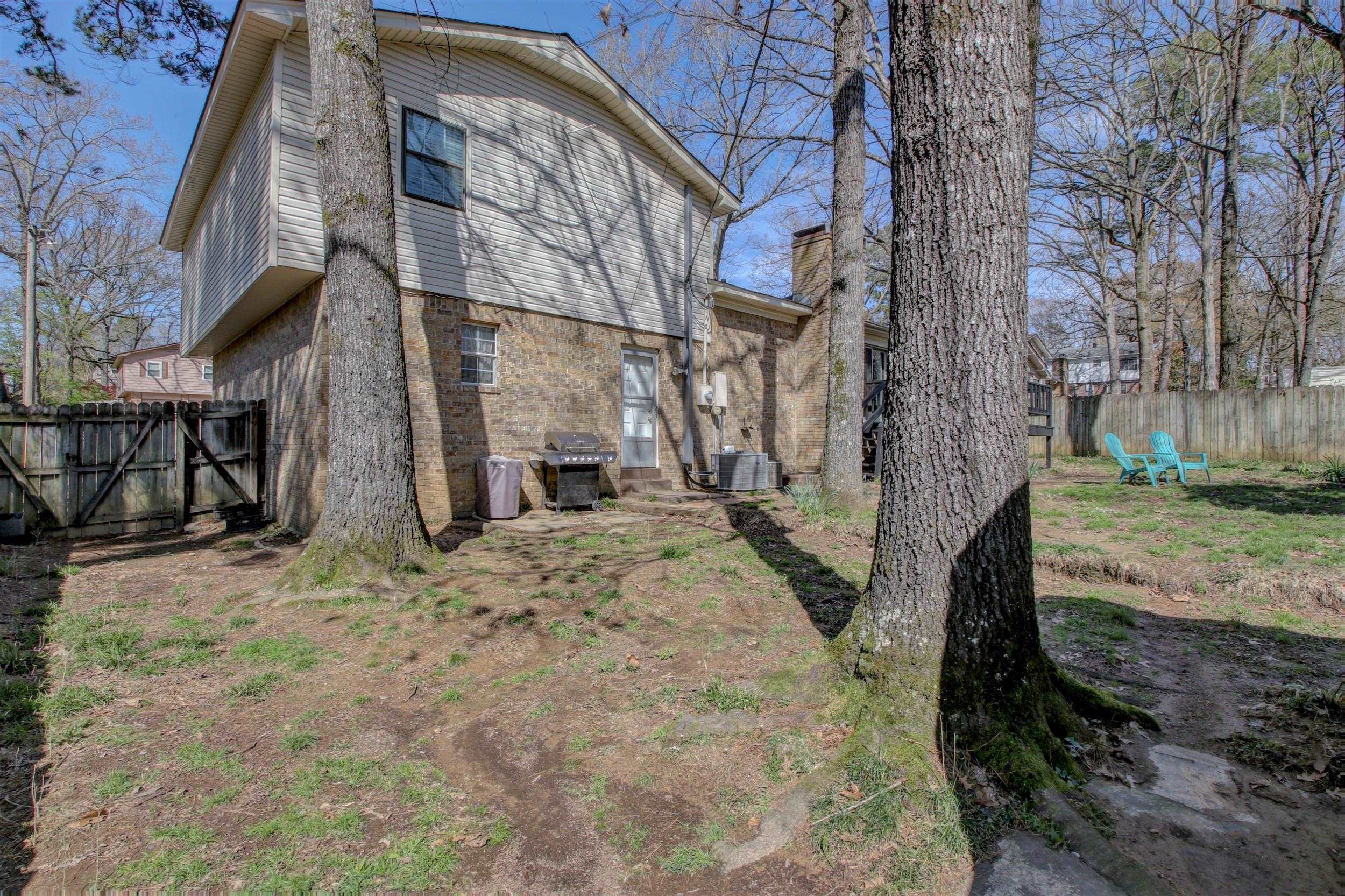 Rear of a two-story brick and gray-sided home, with mature trees and turquoise chairs in the fenced yard.