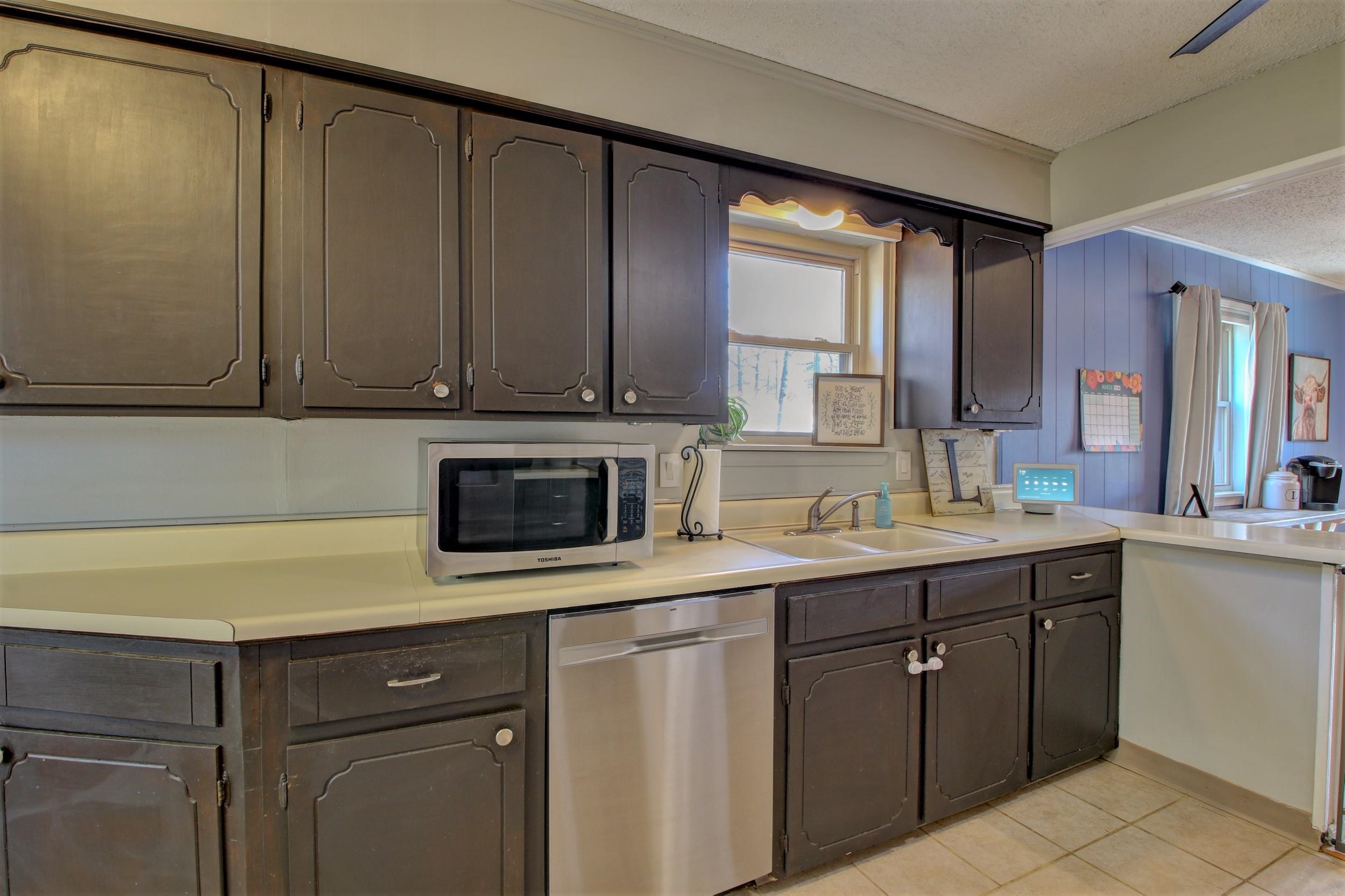 Kitchen with dark cabinets, pale counters, stainless dishwasher, and a double-basin sink beneath a window.