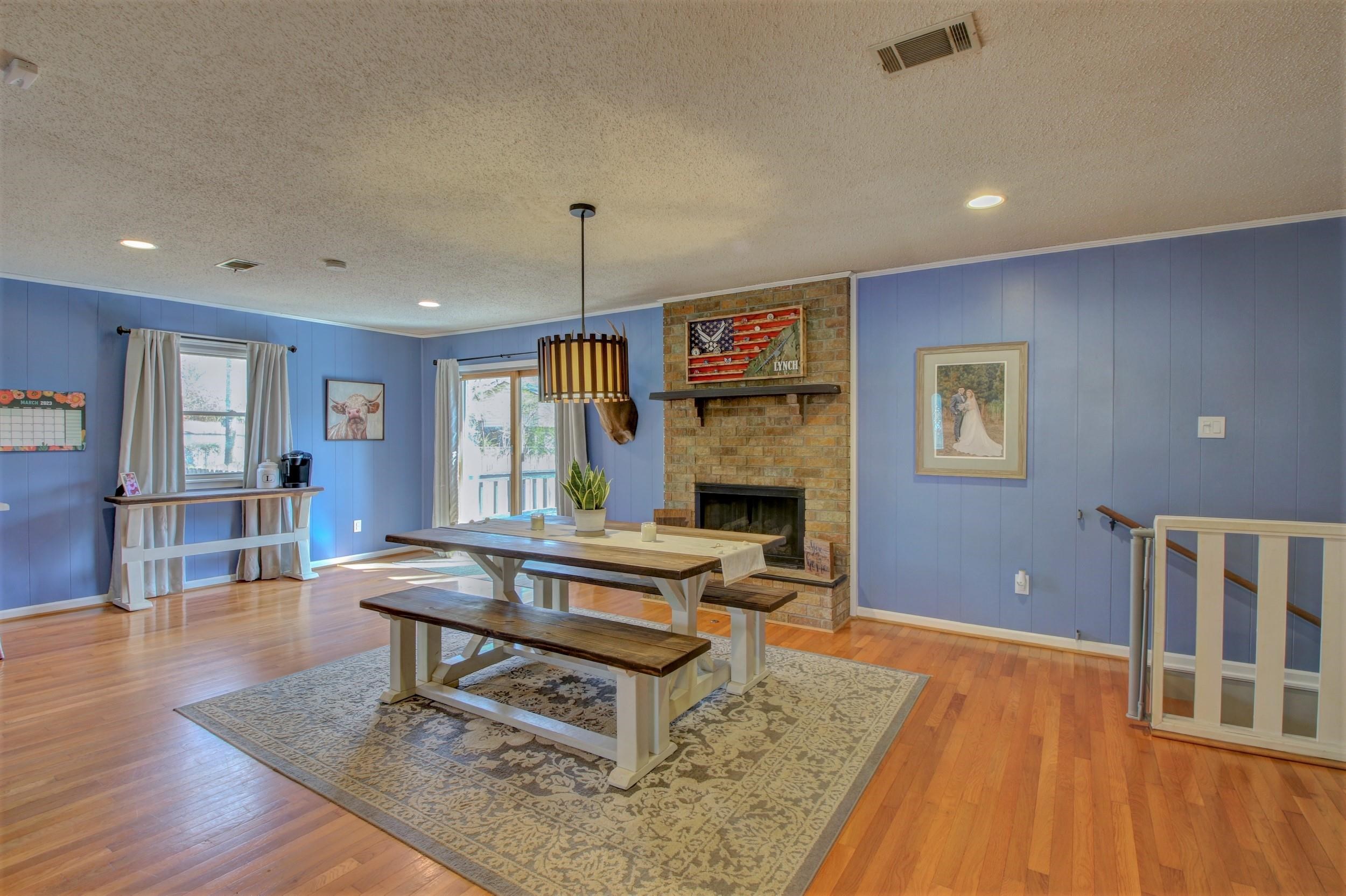 Dining area with blue paneled walls, a brick fireplace, and a farmhouse table on a patterned rug.