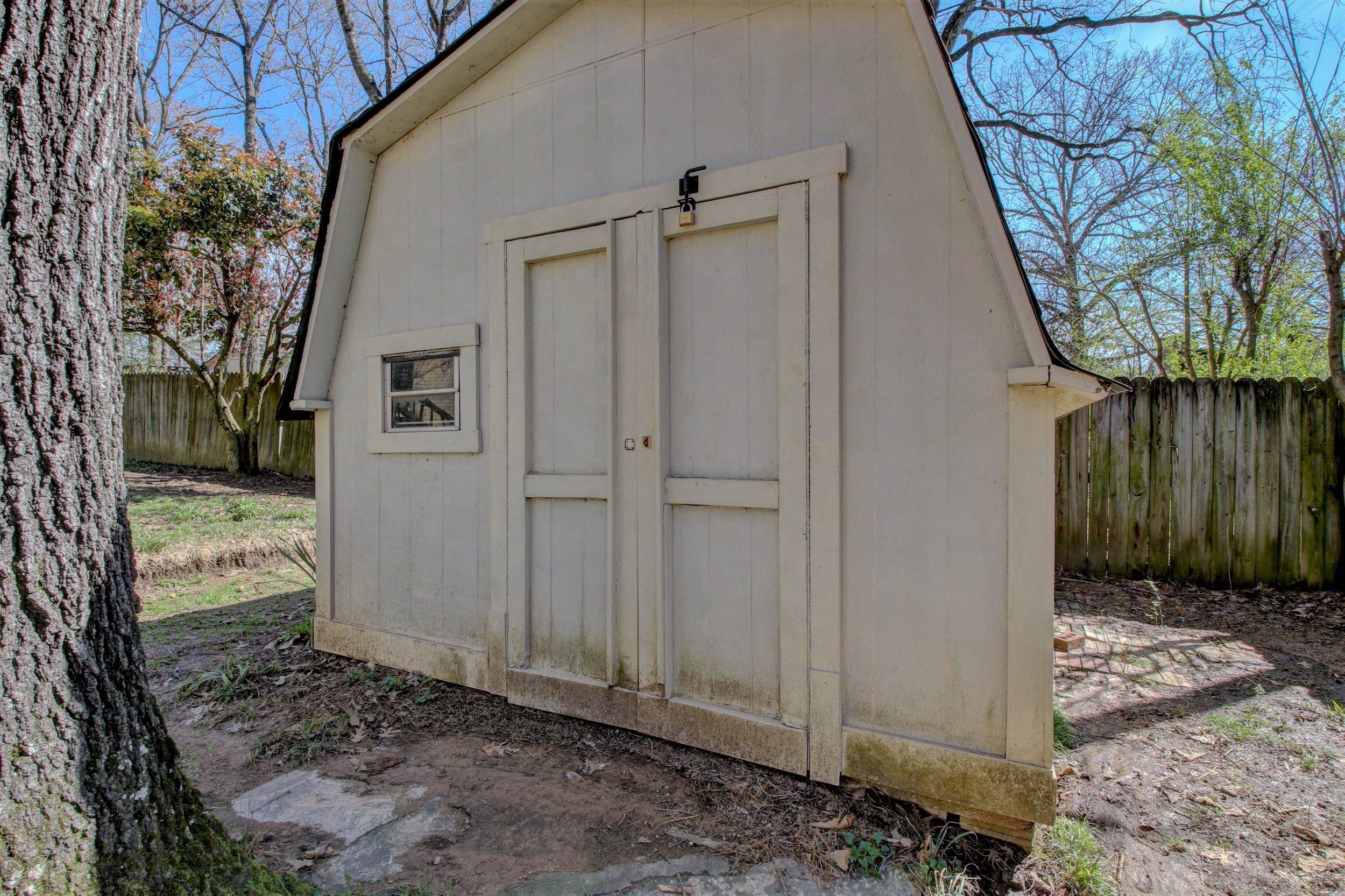 Light-colored backyard shed with a gambrel roof, padlocked double doors, and a small window.