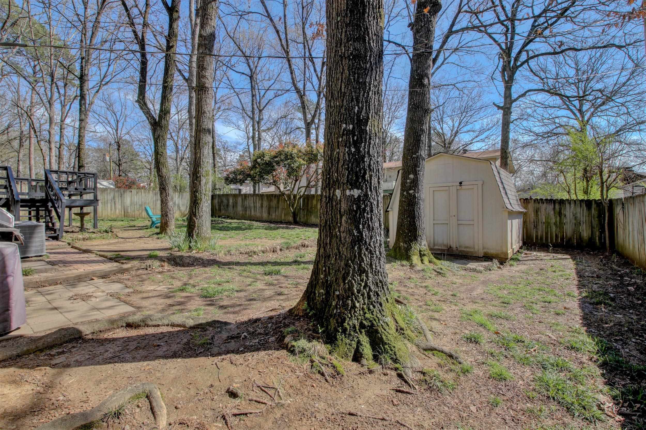 Fenced backyard with mature trees, a raised deck, paved patio, and storage shed.
