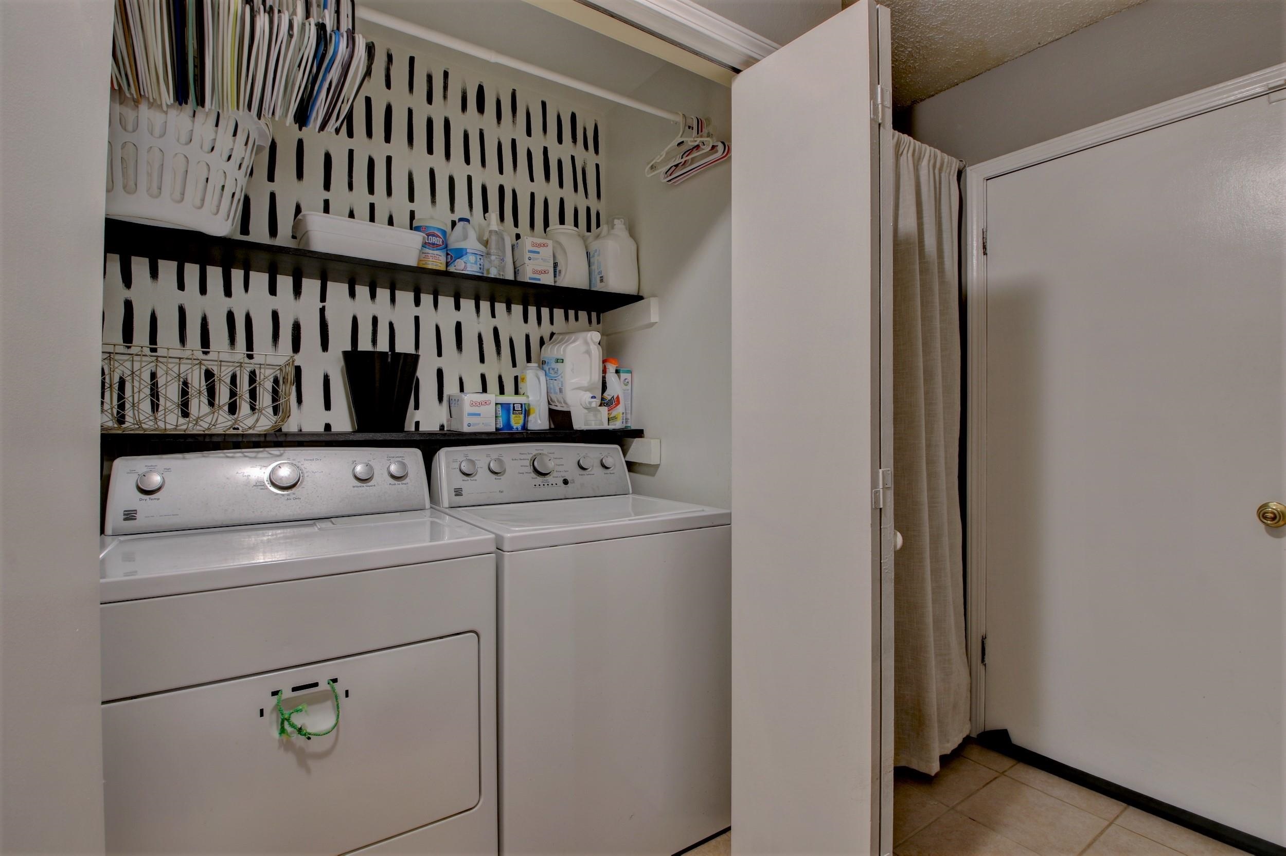 Laundry room with a white washer and dryer beneath storage shelves and a patterned wall.