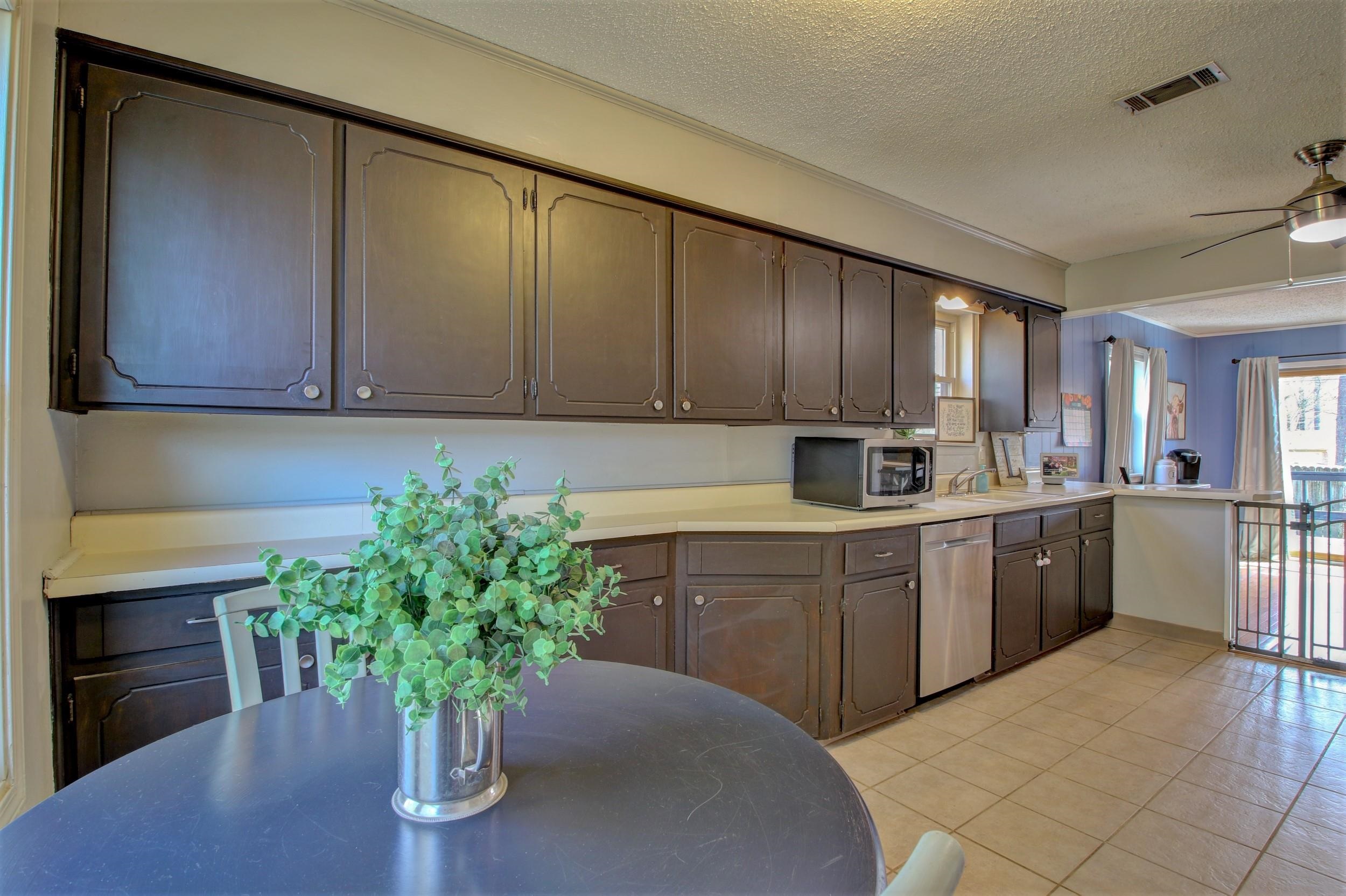 Kitchen with dark cabinets, pale countertops, stainless steel dishwasher, and a round table with a potted plant.