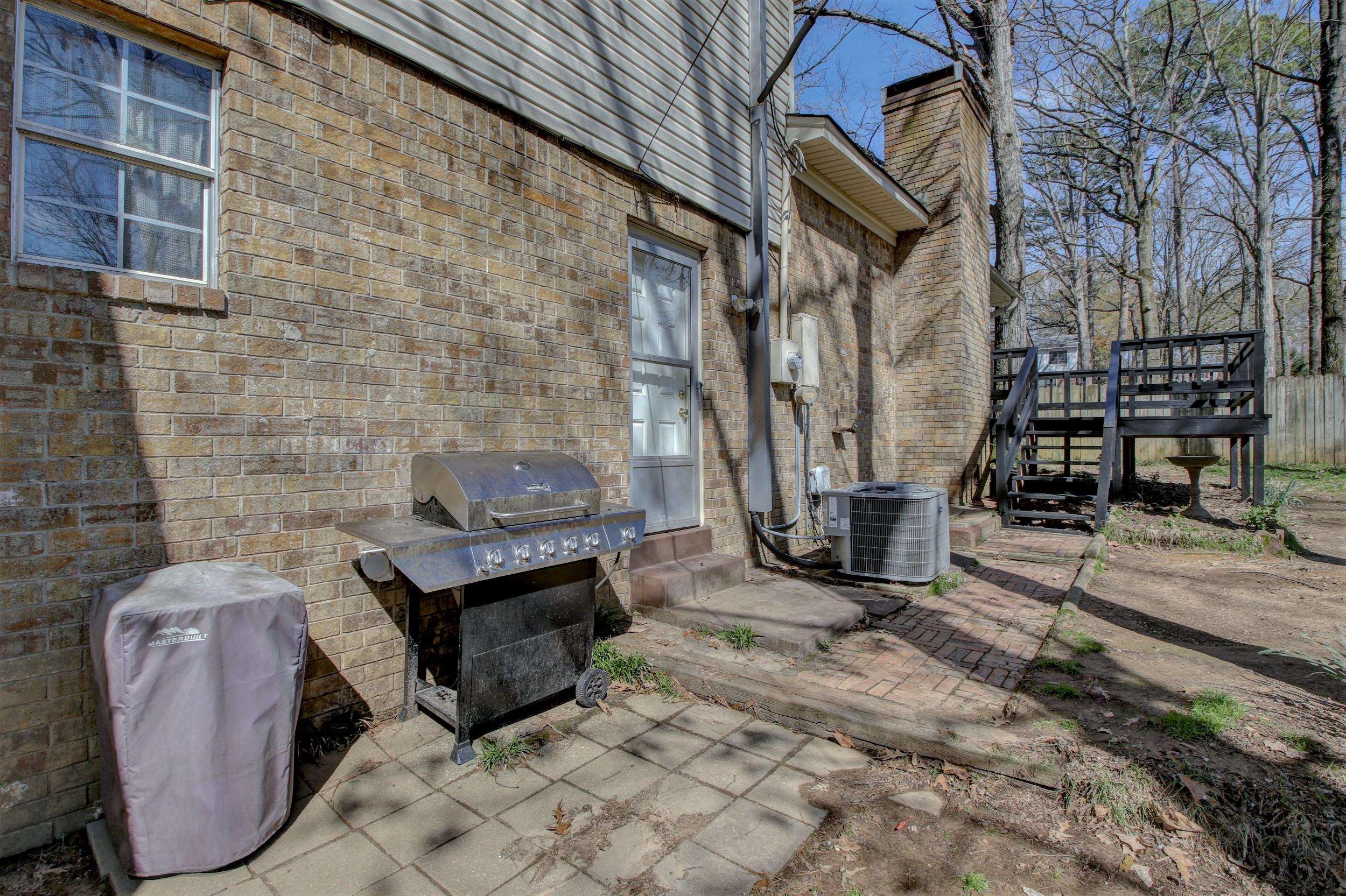 Brick home patio with a covered grill and raised black deck beside a wooded backyard.