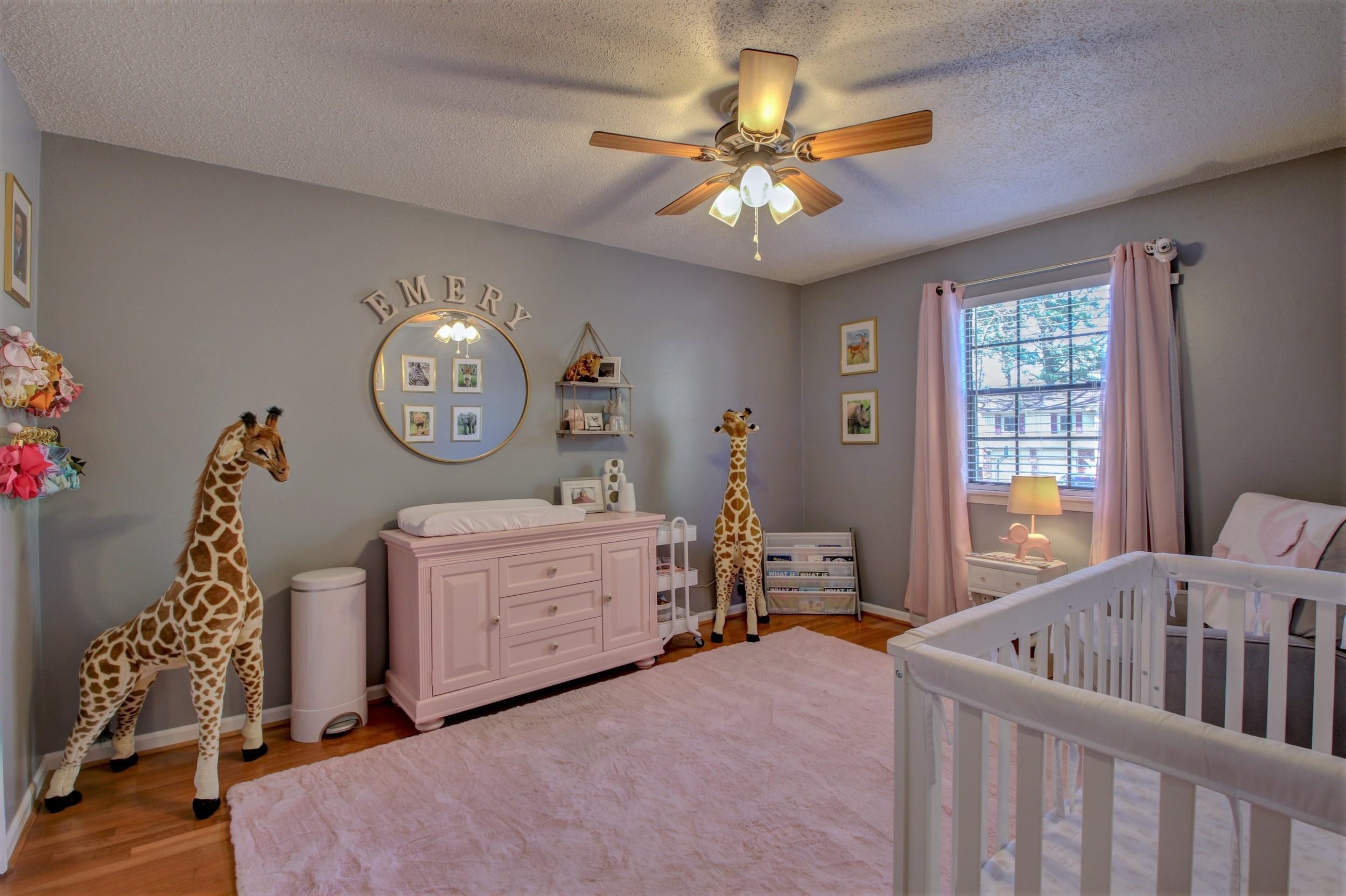Nursery with a white crib, pink furnishings, giraffe decorations, and a round mirror marked “EMERY.”