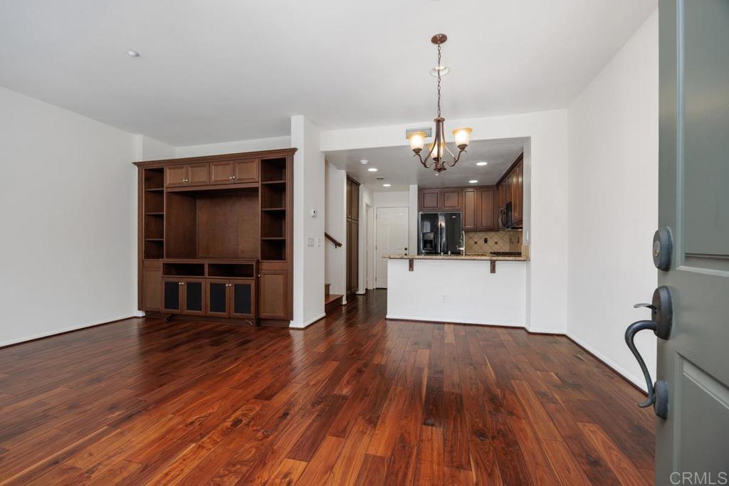 Vacant living room with hardwood floors, built-in wood cabinetry, and a chandelier overlooking the kitchen.
