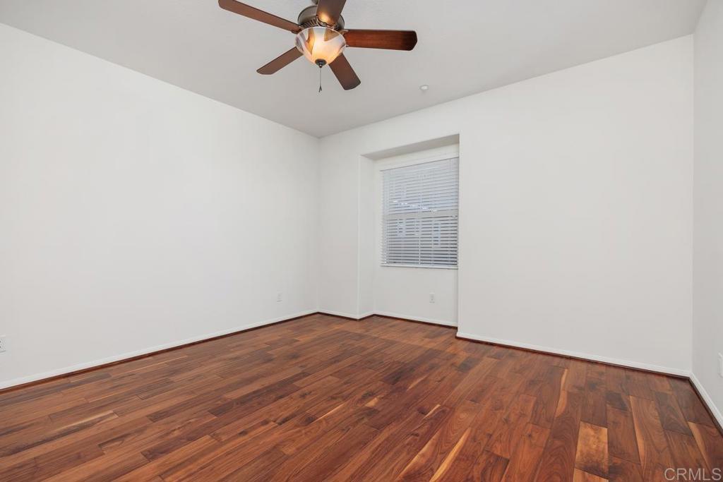 Empty bedroom with reddish-brown hardwood floors, a ceiling fan, and a window with blinds.