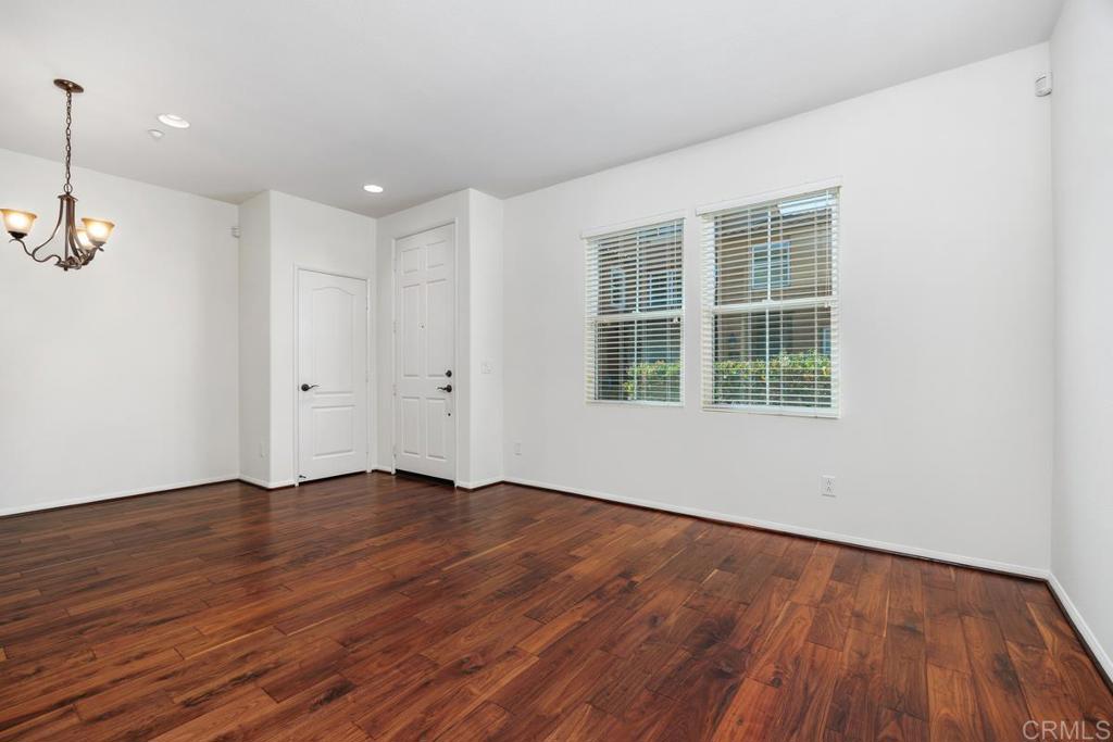 Empty room with reddish-brown hardwood floors, two windows with blinds, and a hanging chandelier.
