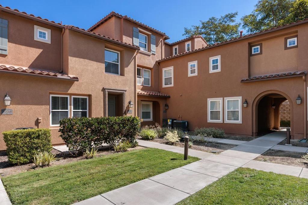 Two-story brown stucco townhouse with red tile roofs and an arched passageway beside a landscaped courtyard.