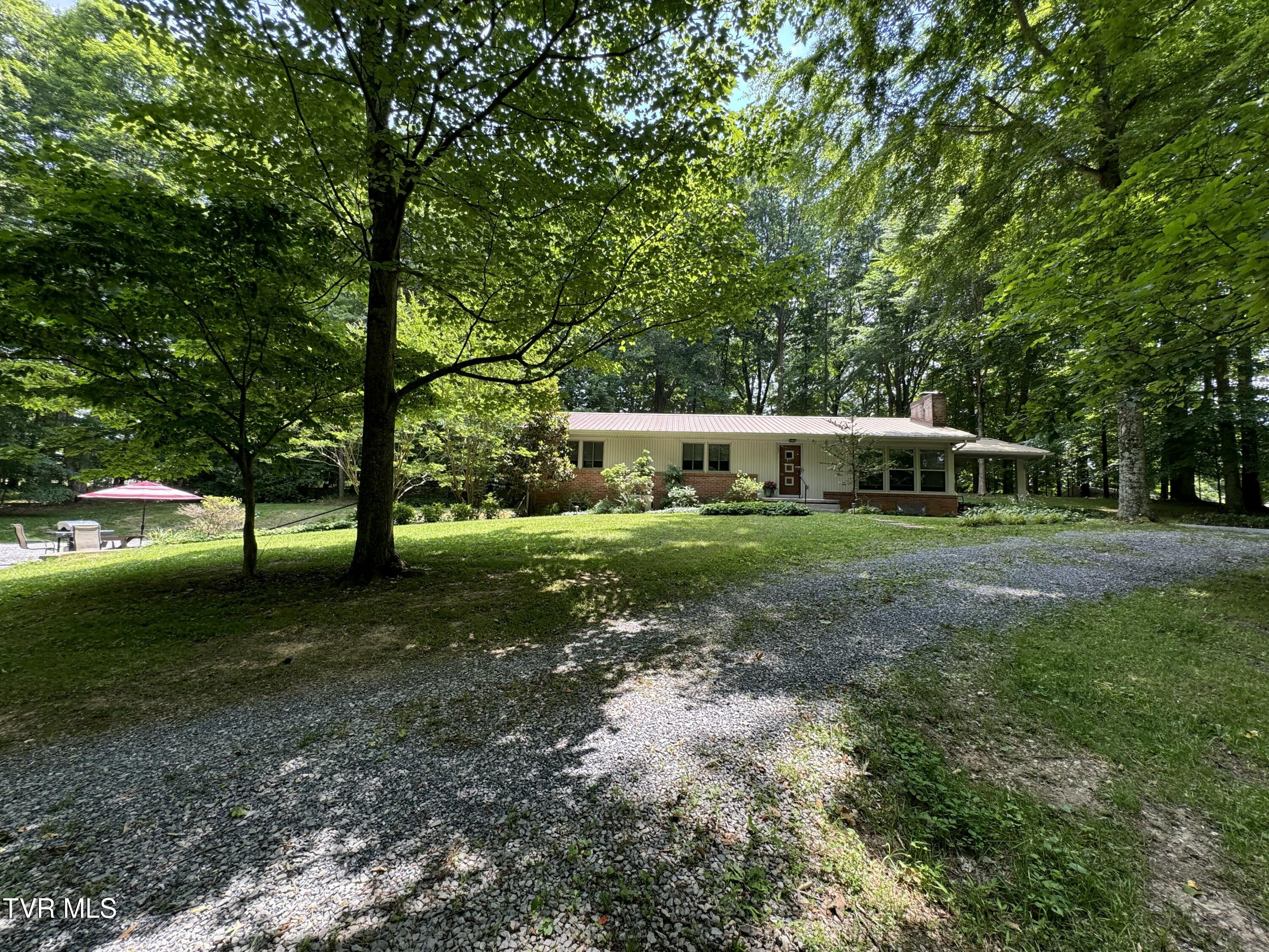 Ranch home nestled among mature trees, with a broad gravel driveway and patio seating.