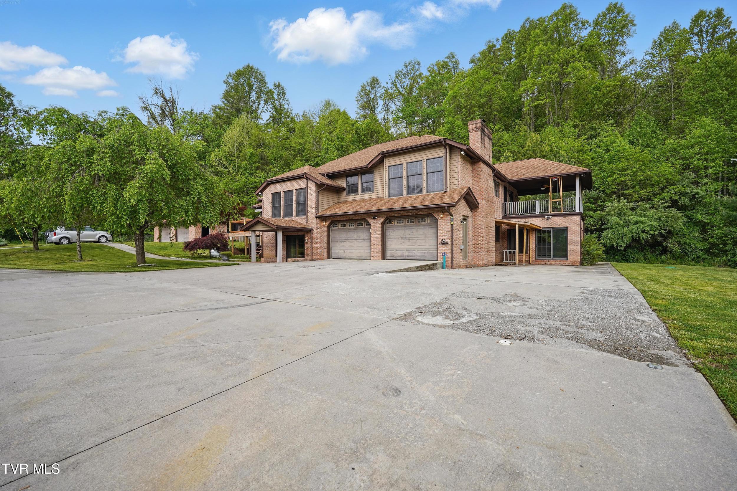 Two-story brick home with a broad driveway, two garage doors, and a covered balcony beside woods.
