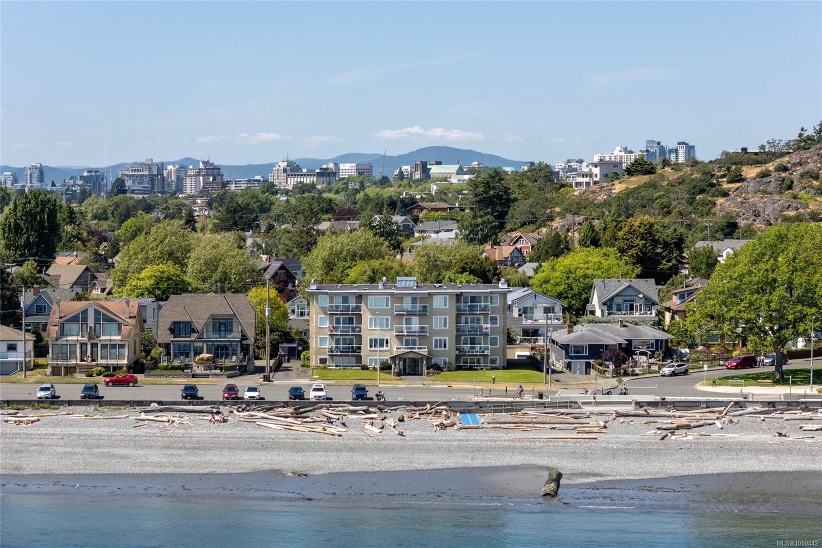 Coastal neighborhood beyond a driftwood-strewn beach, with city buildings and mountains in the distance.