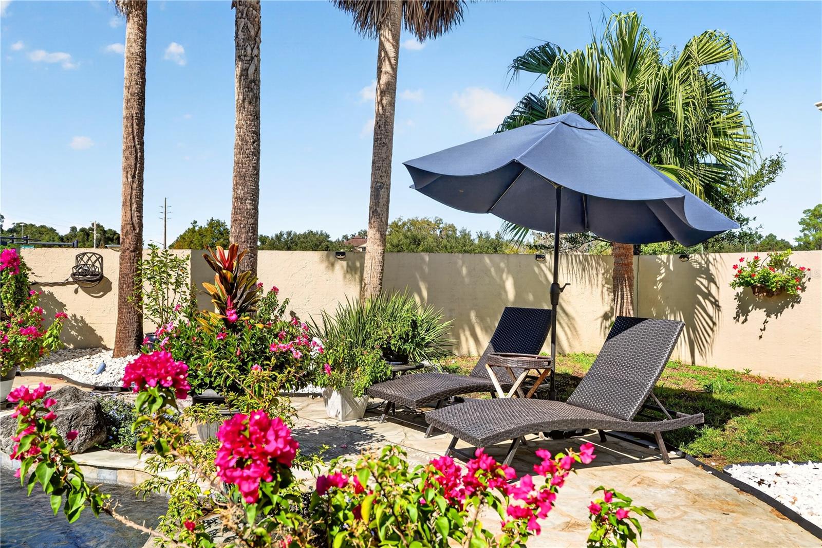 Stone poolside patio with chaise lounges beneath an umbrella, surrounded by palms and flowering plants.
