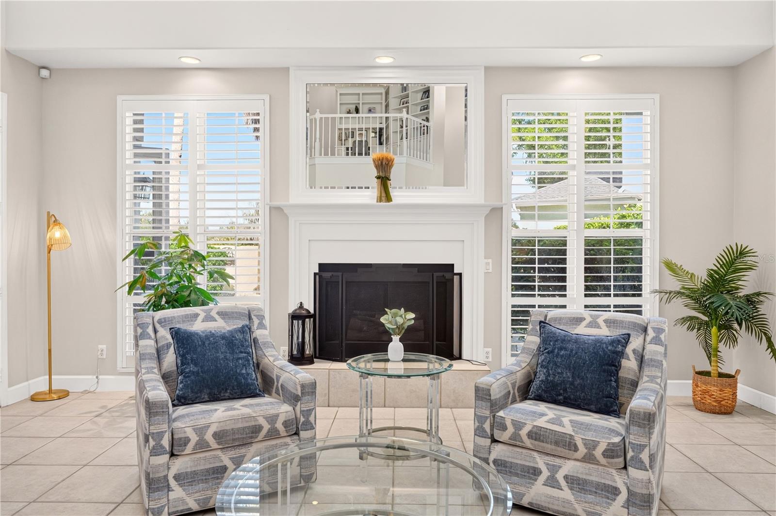 Living room with a white fireplace, shuttered windows, patterned armchairs, and a glass coffee table.