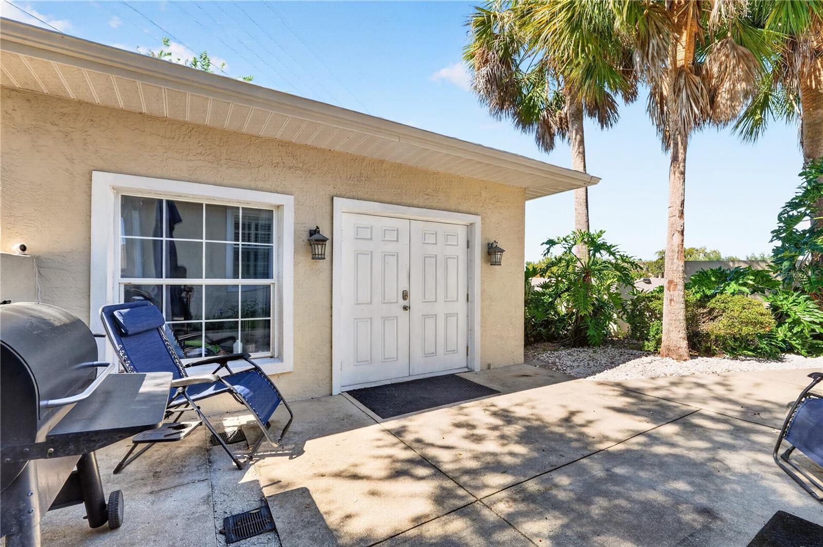 Concrete patio with white double doors, palm trees, outdoor chairs, and a barbecue grill.