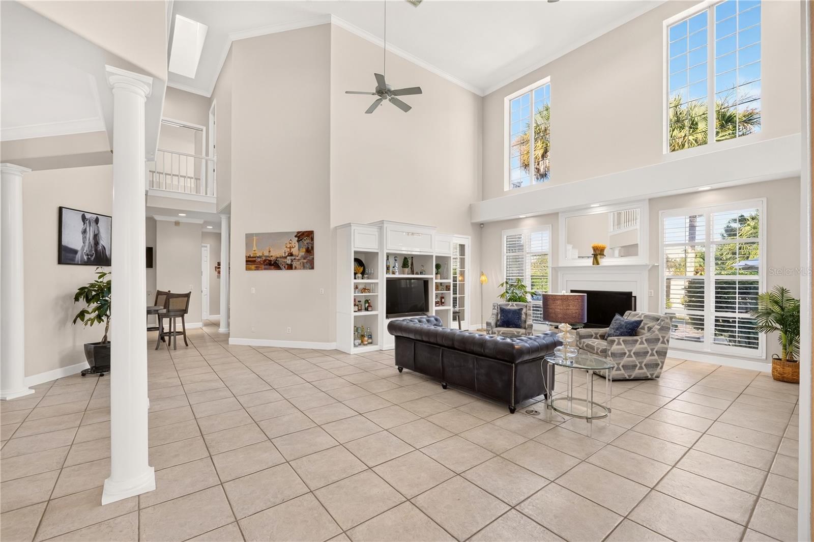 Double-height living room with tile floors, built-in TV shelving, a fireplace, and large windows.