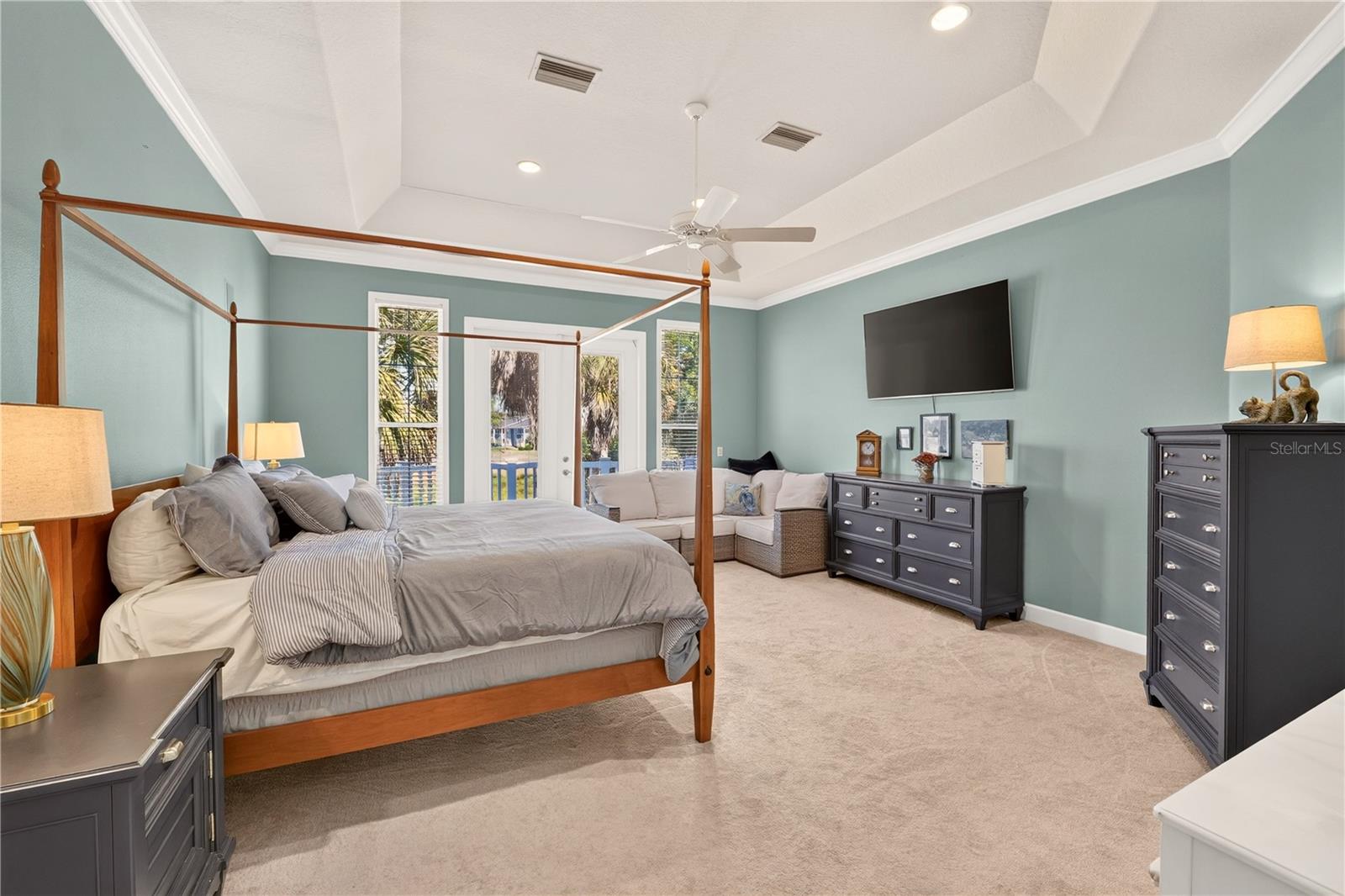 Furnished bedroom with a four-poster bed, tray ceiling, and sitting area beside glass doors.