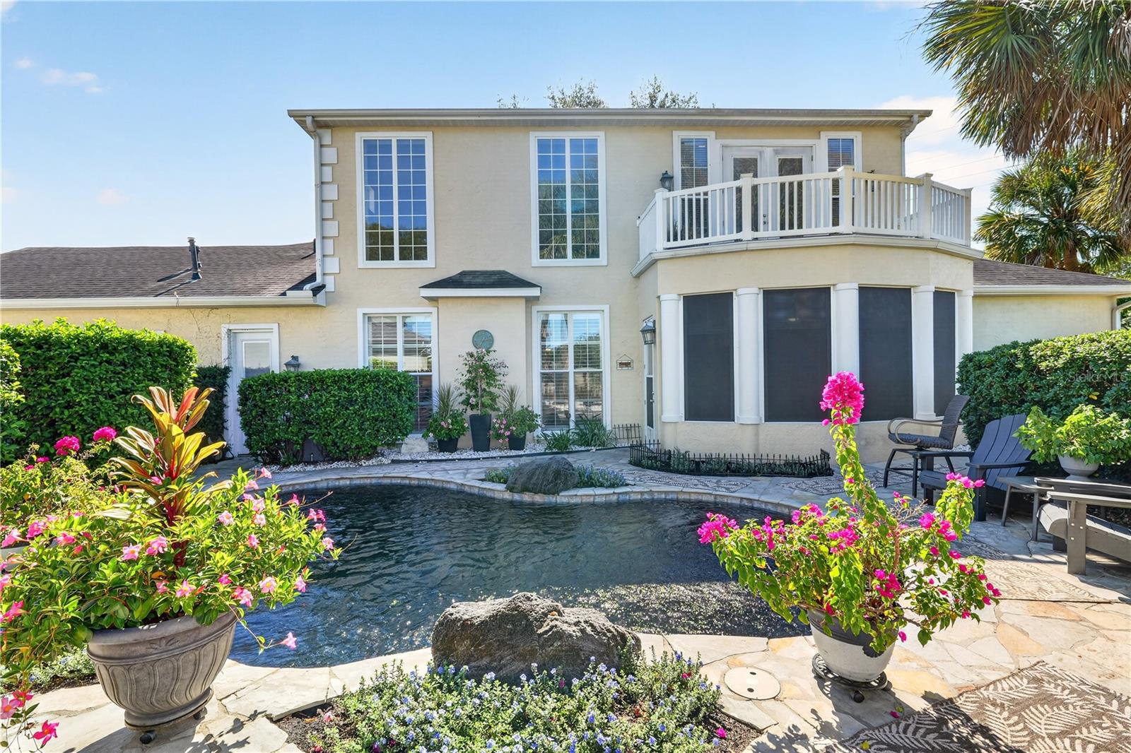 Home’s rear exterior with a curved pool, upper balcony, stone patio, and flowering plants.