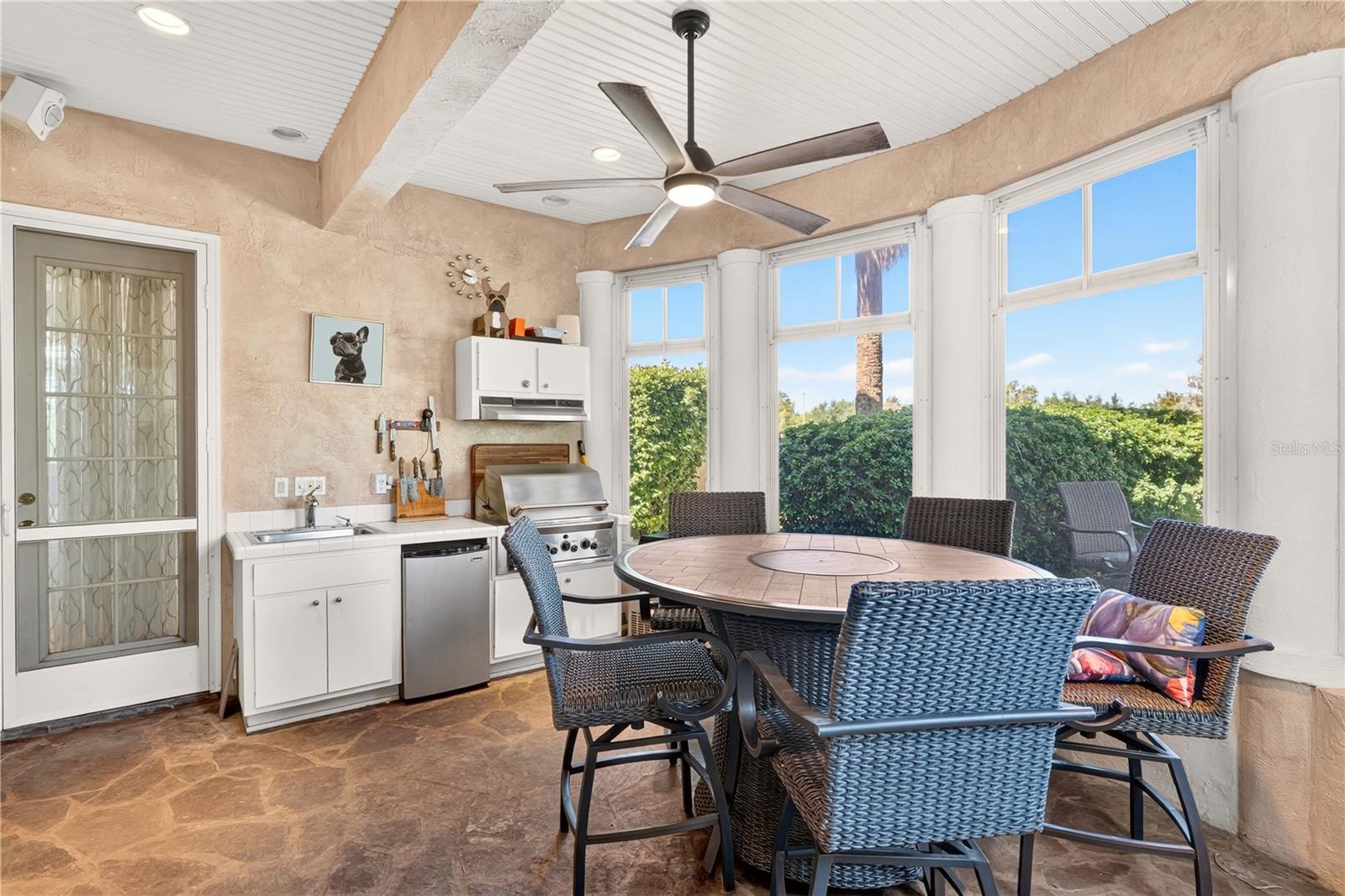 Covered patio with wicker dining set, built-in grill, and large windows overlooking hedges.