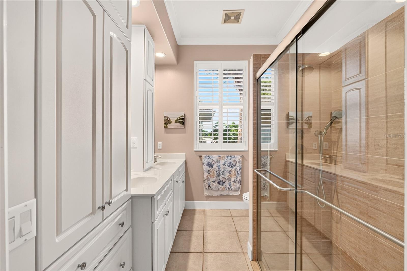 Bathroom with double vanity and plantation shutters beside a glass shower with tan tile.