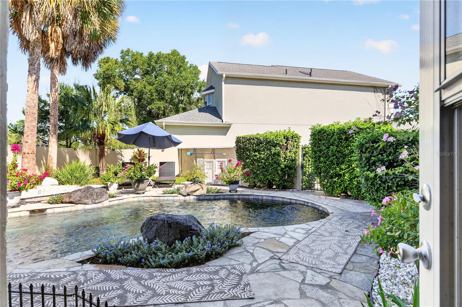 Backyard pool with a stone patio, umbrella seating, and tropical landscaping.