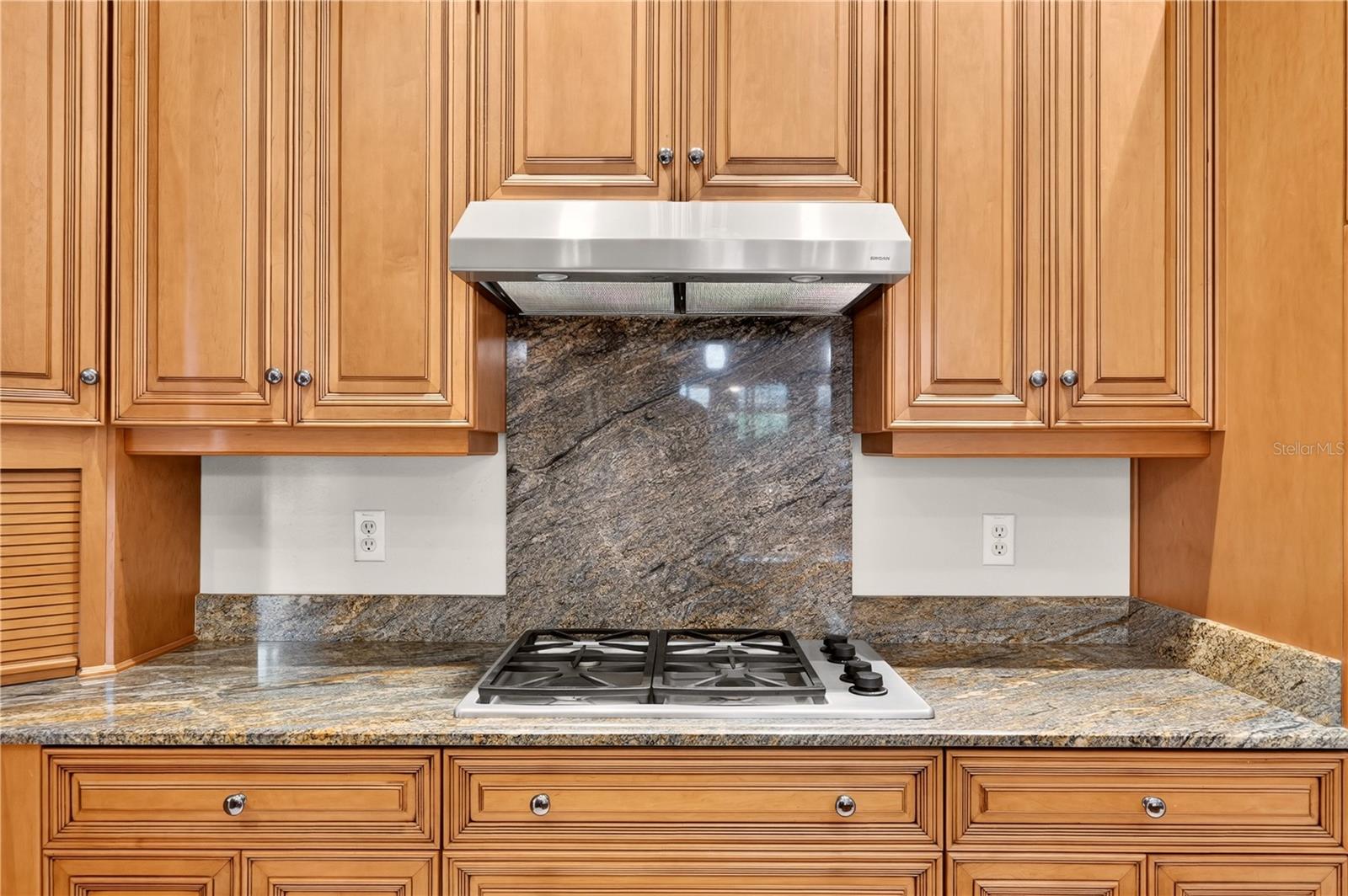 Kitchen cooktop beneath a stainless range hood, framed by wood cabinets and patterned stone surfaces.