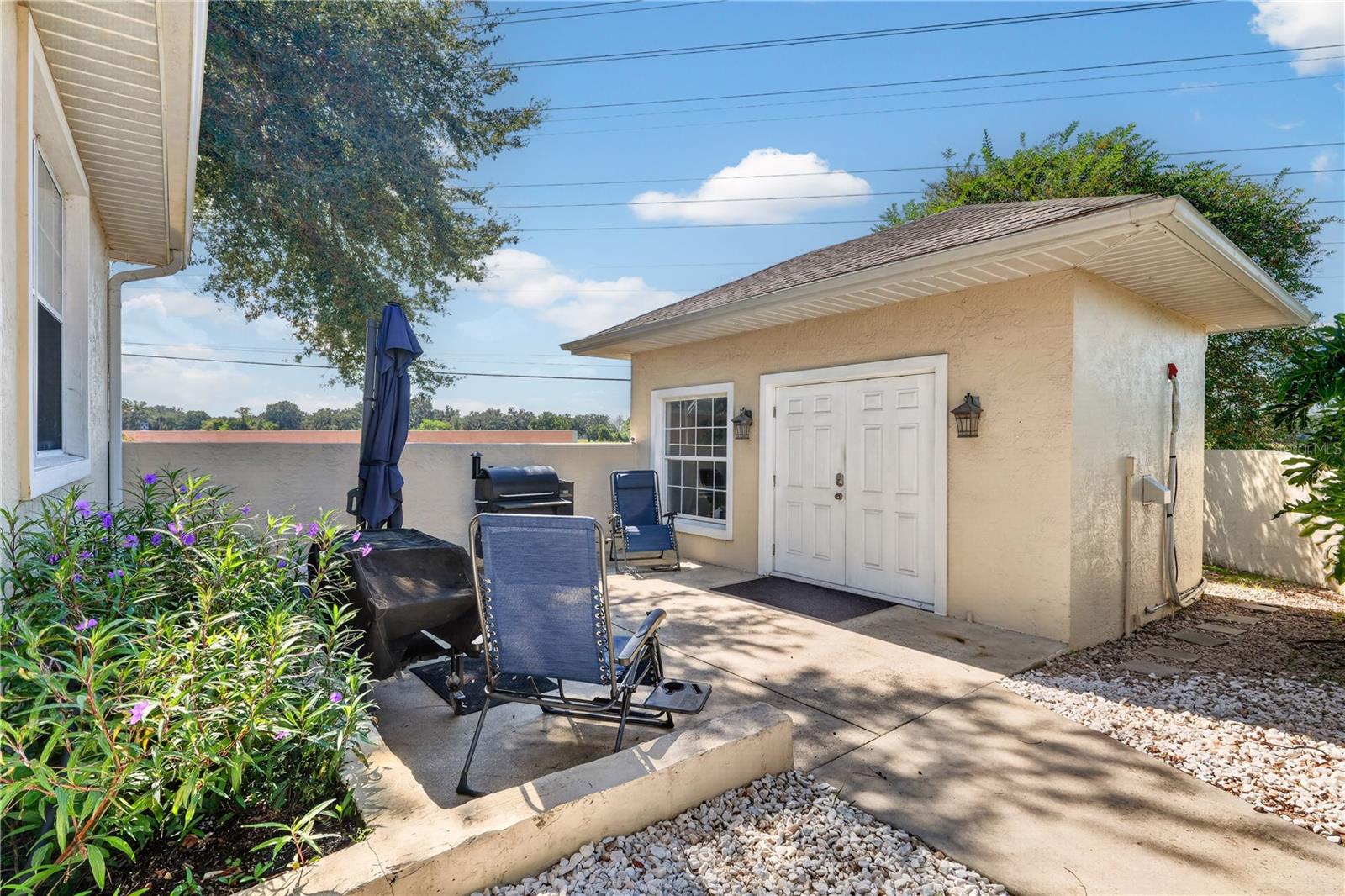 Paved patio with outdoor seating, barbecue grill, flowering plants, and a small stucco outbuilding.