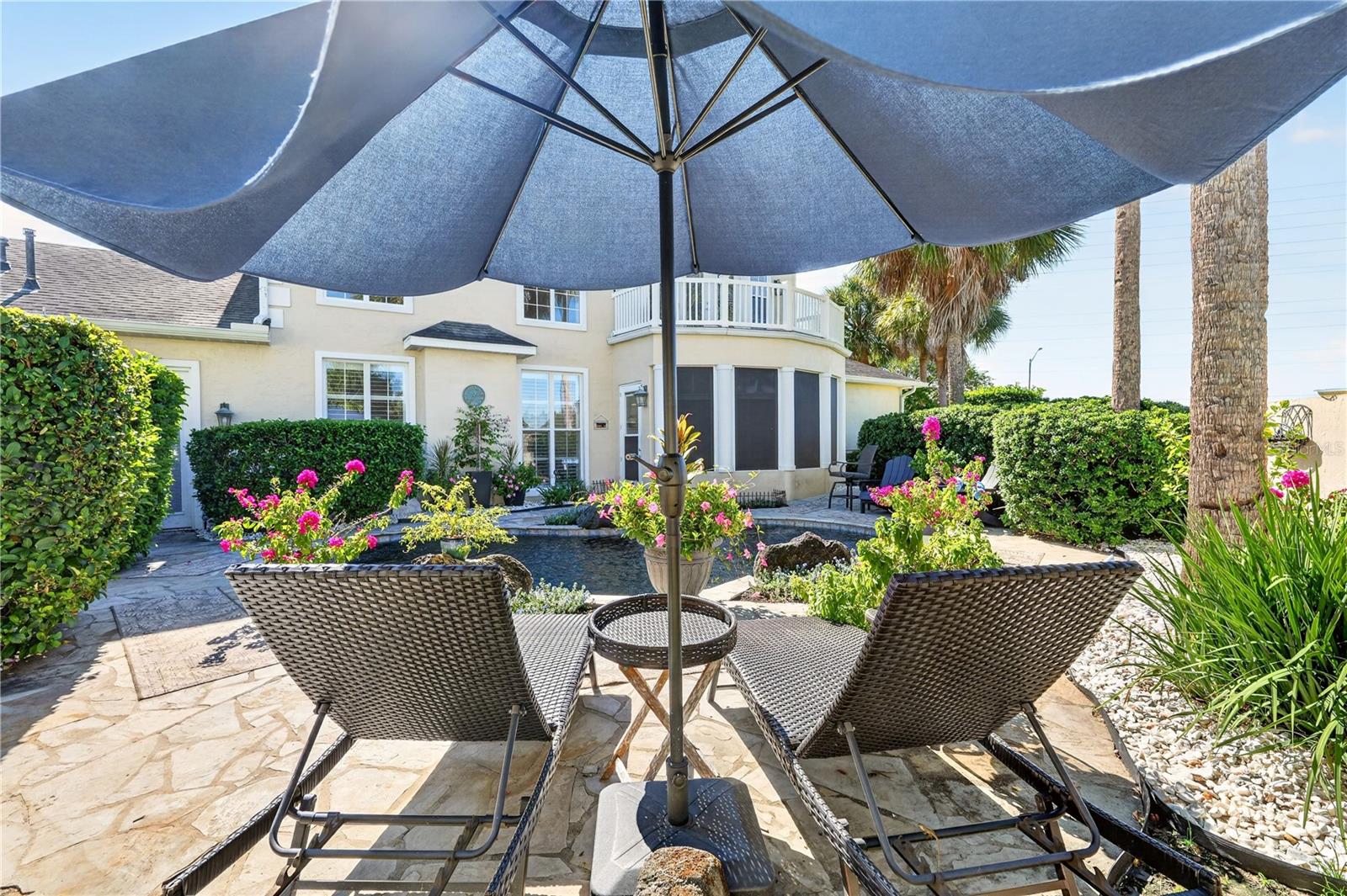 Backyard patio with woven lounge chairs, a blue umbrella, flowering beds, and a small water feature.