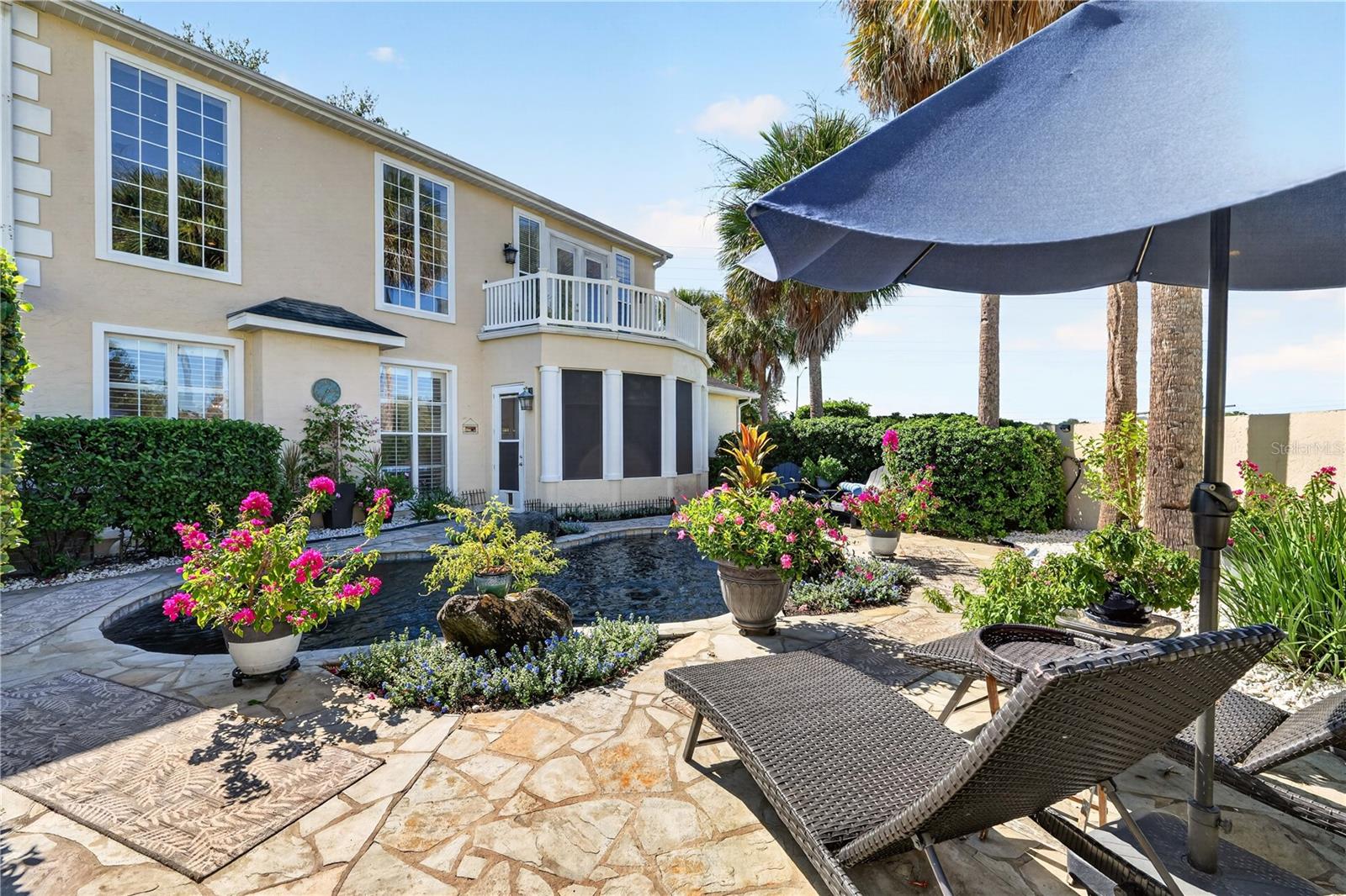 Two-story home beside a landscaped pond and stone patio with lounge chairs under an umbrella.